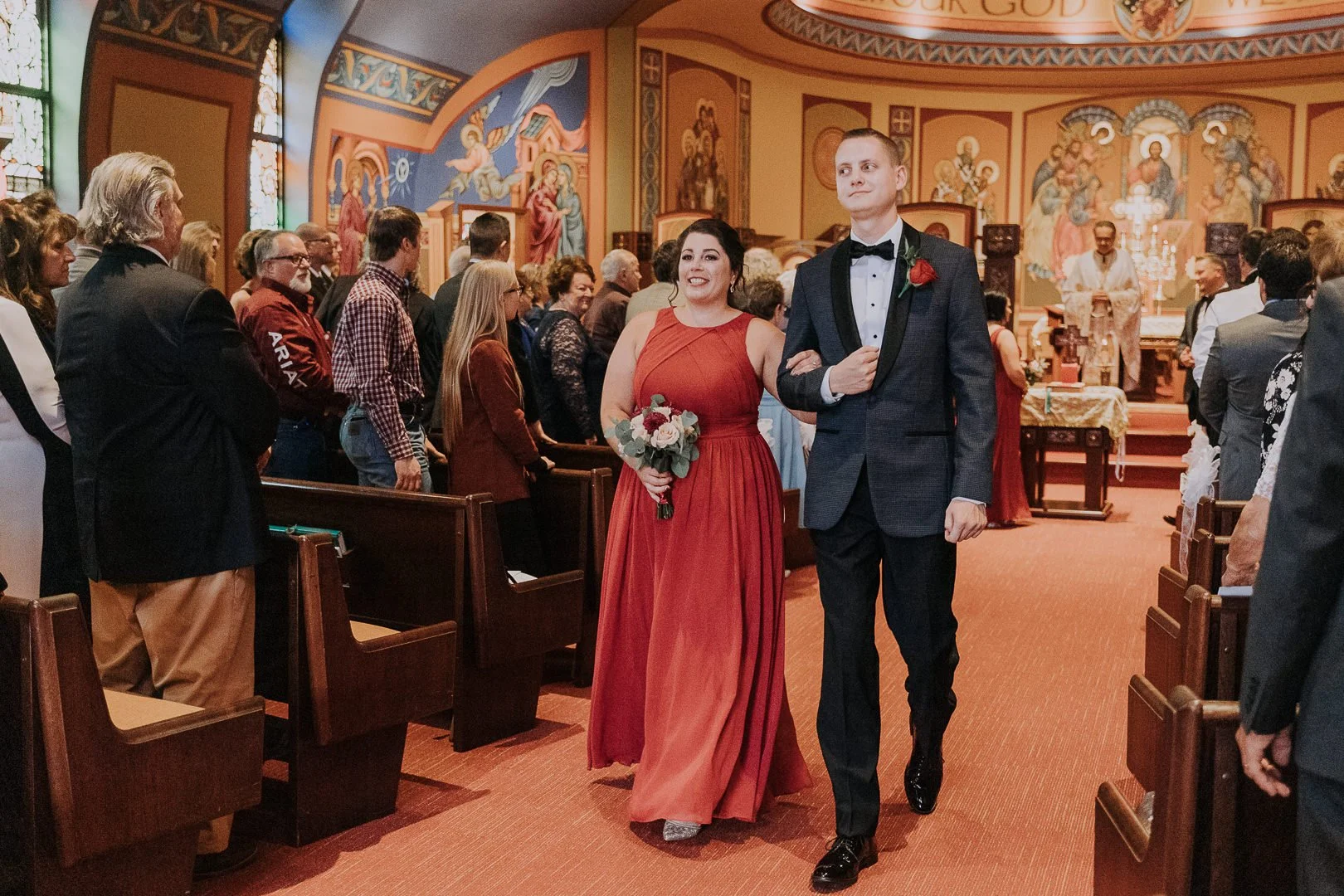 A bride and groom walk down the aisle inside a church filled with guests attending a wedding ceremony. The bride is wearing a red dress and holding a bouquet, and the groom is dressed in a dark tuxedo with a red boutonniere.