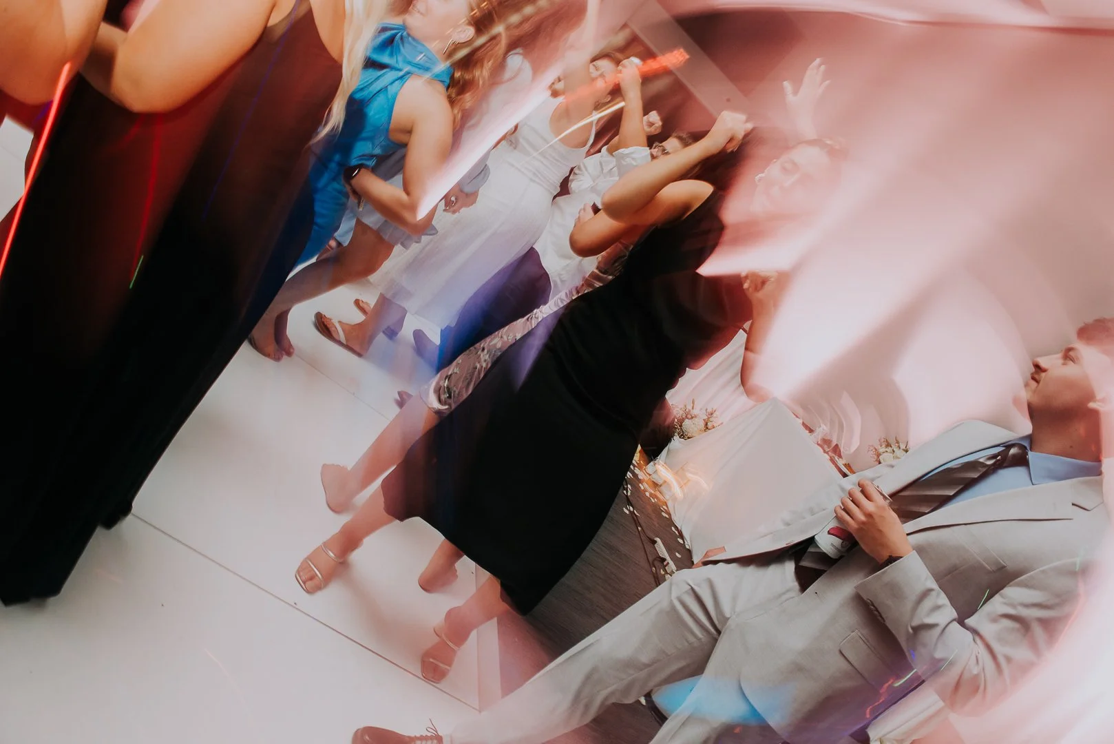A group of people dancing and socializing at a party or celebration, with a woman in a black dress and a man in a light gray suit in the foreground.