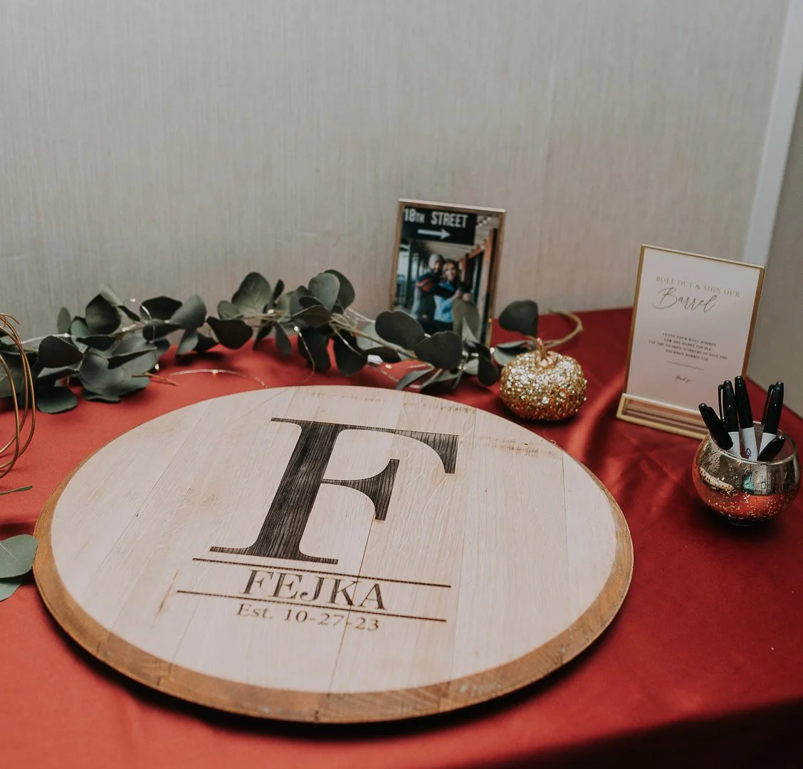 Wedding guest book display with a large wooden initial 'F' and the name 'Fejka' along with the date '10-27-23', decorated with greenery, a framed photo, a glittery pumpkin, a sign to sign the guest book, and pens in a silver cup, on a red tablecloth.