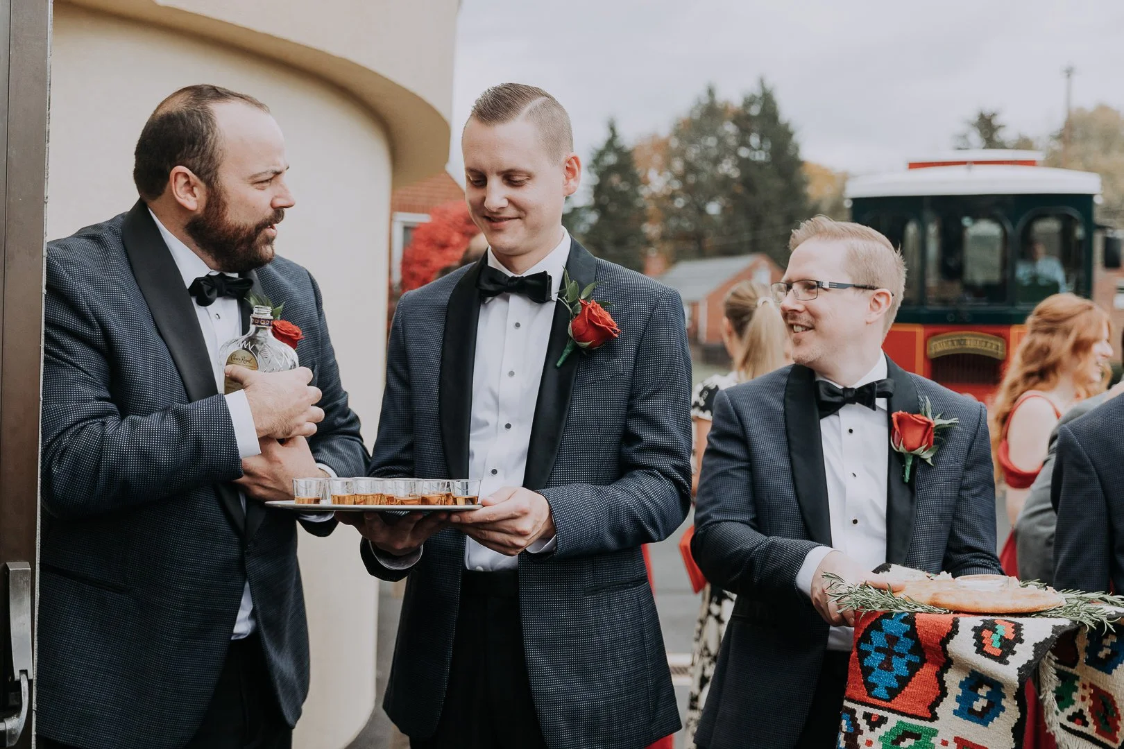 Three men in tuxedos with red roses on their lapels, two of them holding trays with drinks and food, standing outdoors at a wedding or celebration.