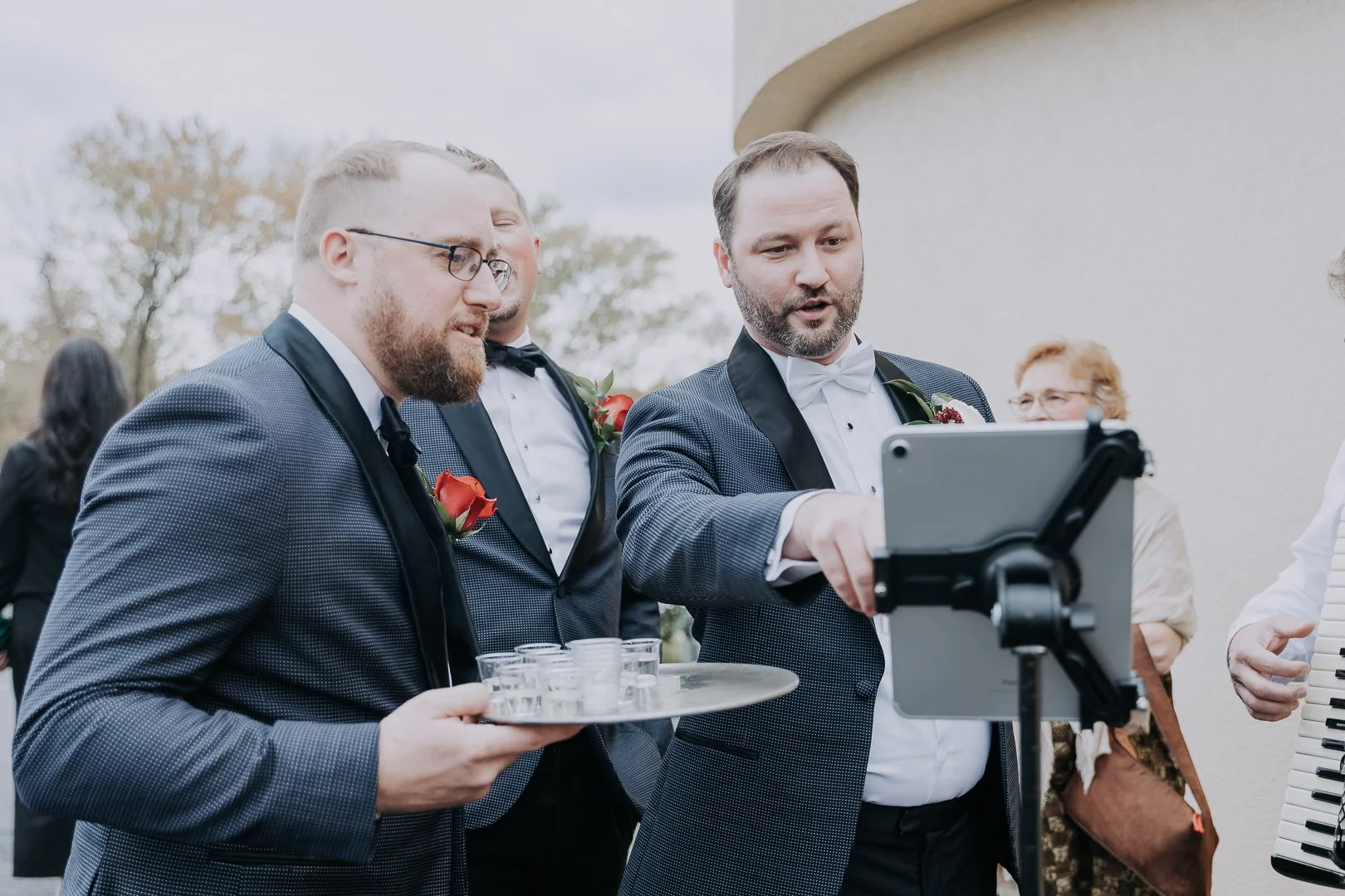 Three men in tuxedos with boutonnières gathered outdoors, looking at a tablet held by one of them, with glasses and a tray of shot glasses.