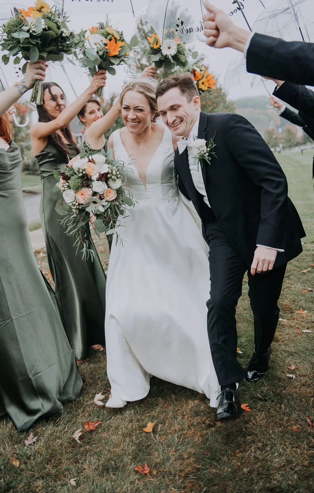 Bride and groom smiling as they walk under umbrellas held by bridesmaids, holding bouquets, during a wedding celebration outdoors in fall.