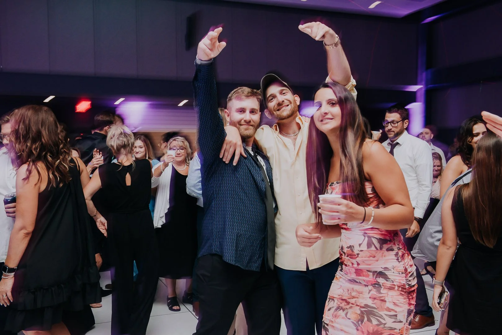 People dancing and socializing at a party with purple lighting, including three individuals in the foreground smiling and posing for the camera, with other guests in the background.