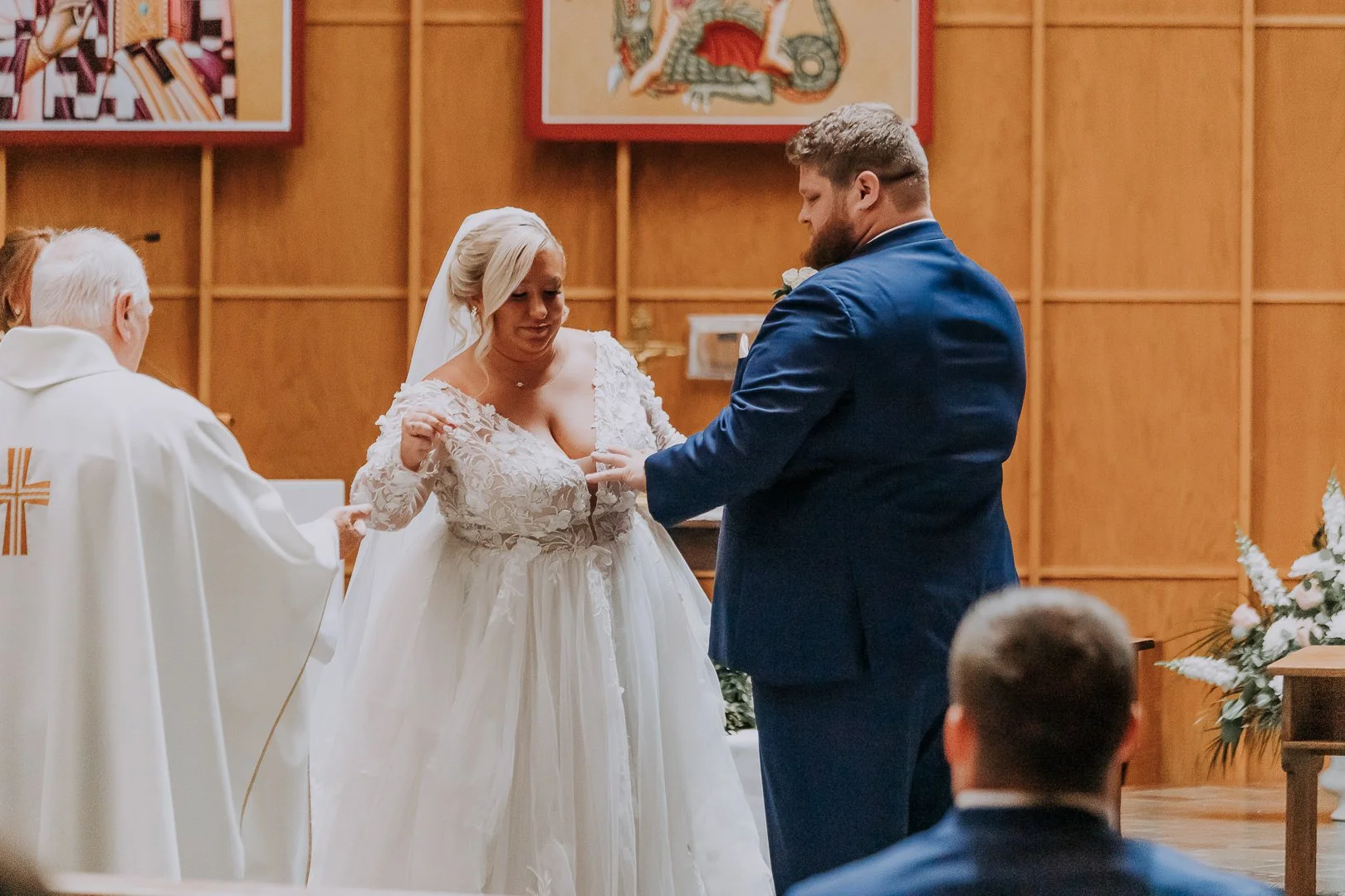 A bride and groom exchanging wedding rings during a church ceremony, with a priest assisting, in a wooden-paneled church with religious artwork in the background.