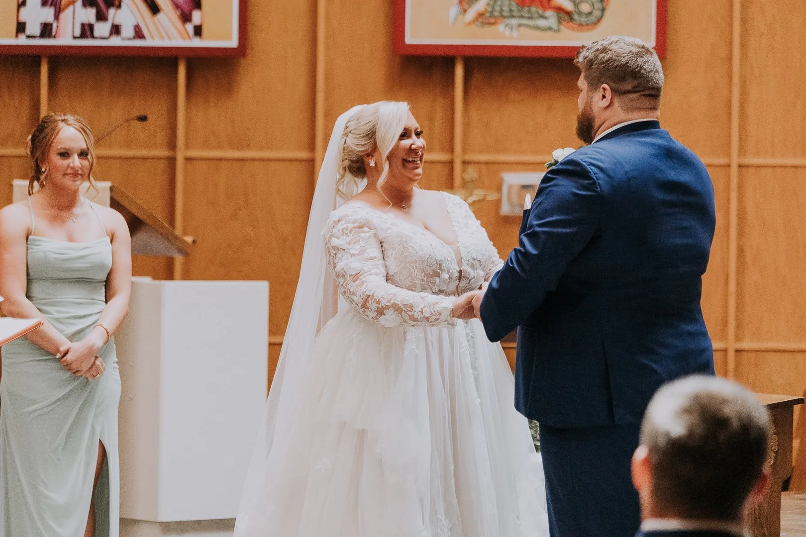 A bride and groom holding hands and smiling during their wedding ceremony, with a bridesmaid standing to the left in a light green dress, set indoors with wooden panel walls.