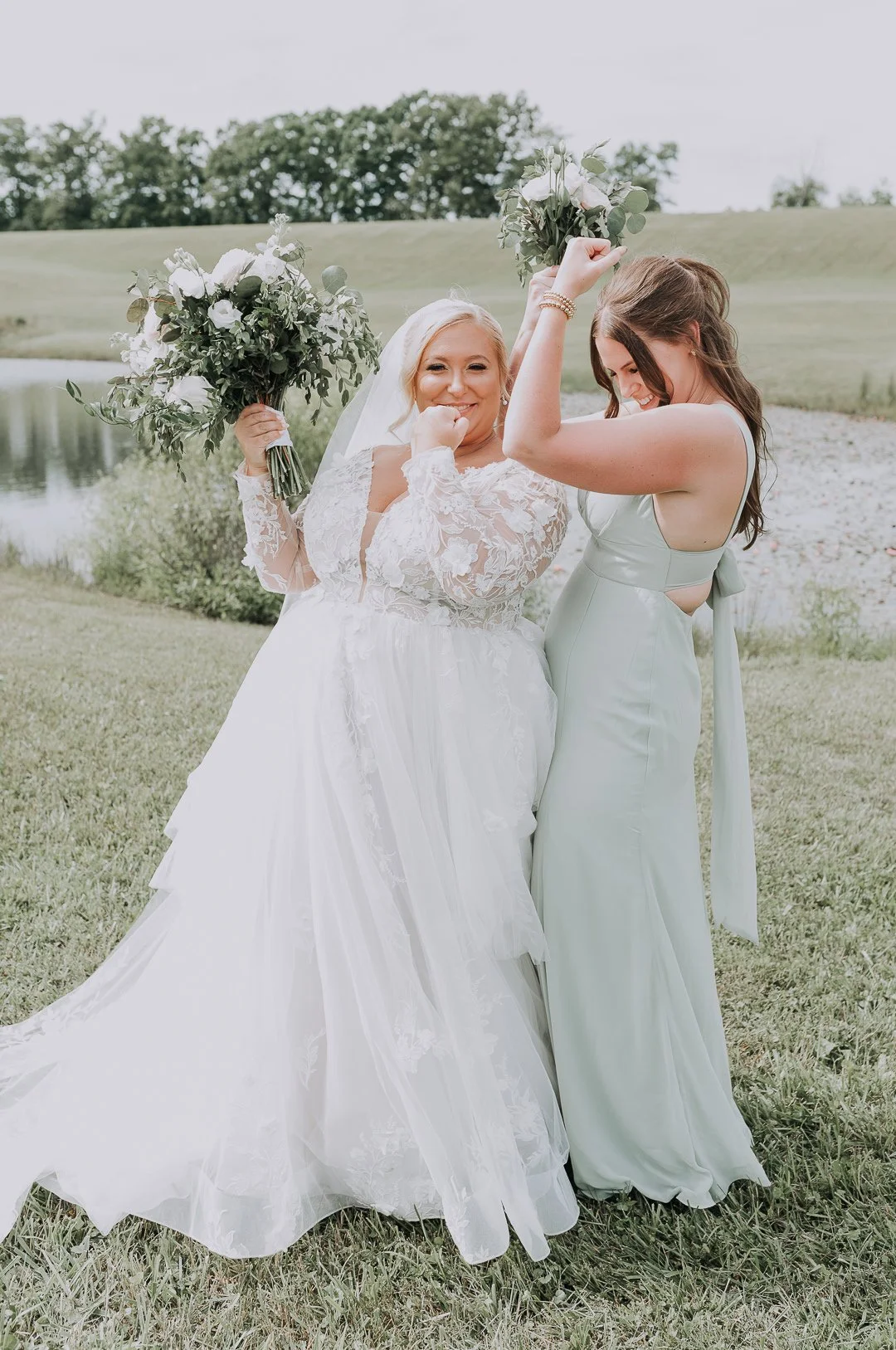 A bride and bridesmaid celebrating outdoors near a lake, holding bouquets, on a grassy area with hills in the background.