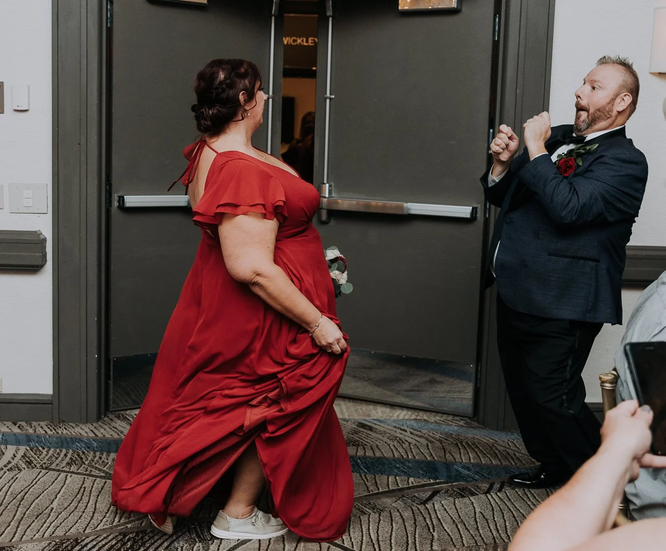 A woman in a red dress and a man in a tuxedo dance happily at a wedding reception, with a dark door in the background and a patterned carpet.