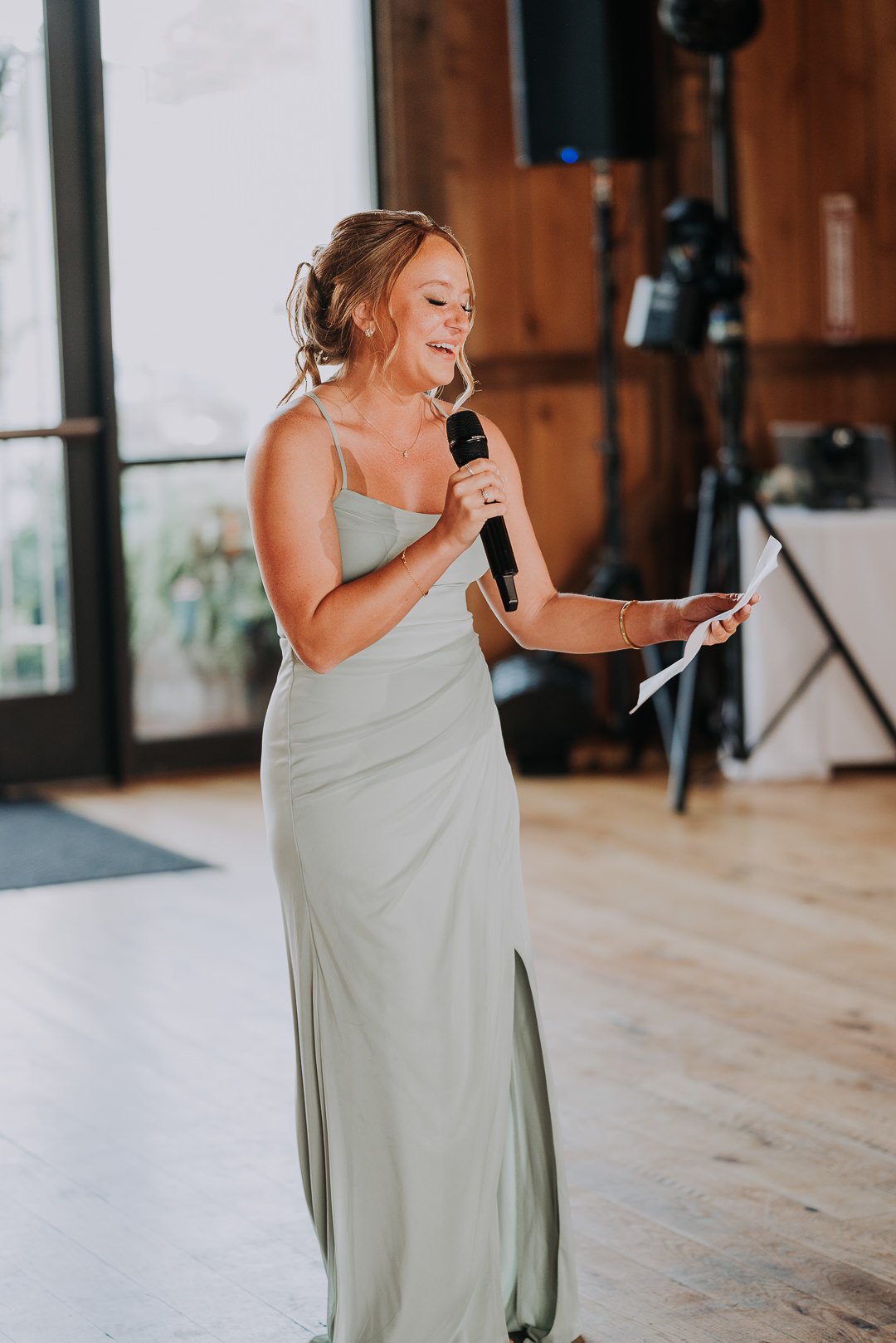 A woman in a light grey dress giving a speech at an indoor event, holding a microphone and reading from a paper.