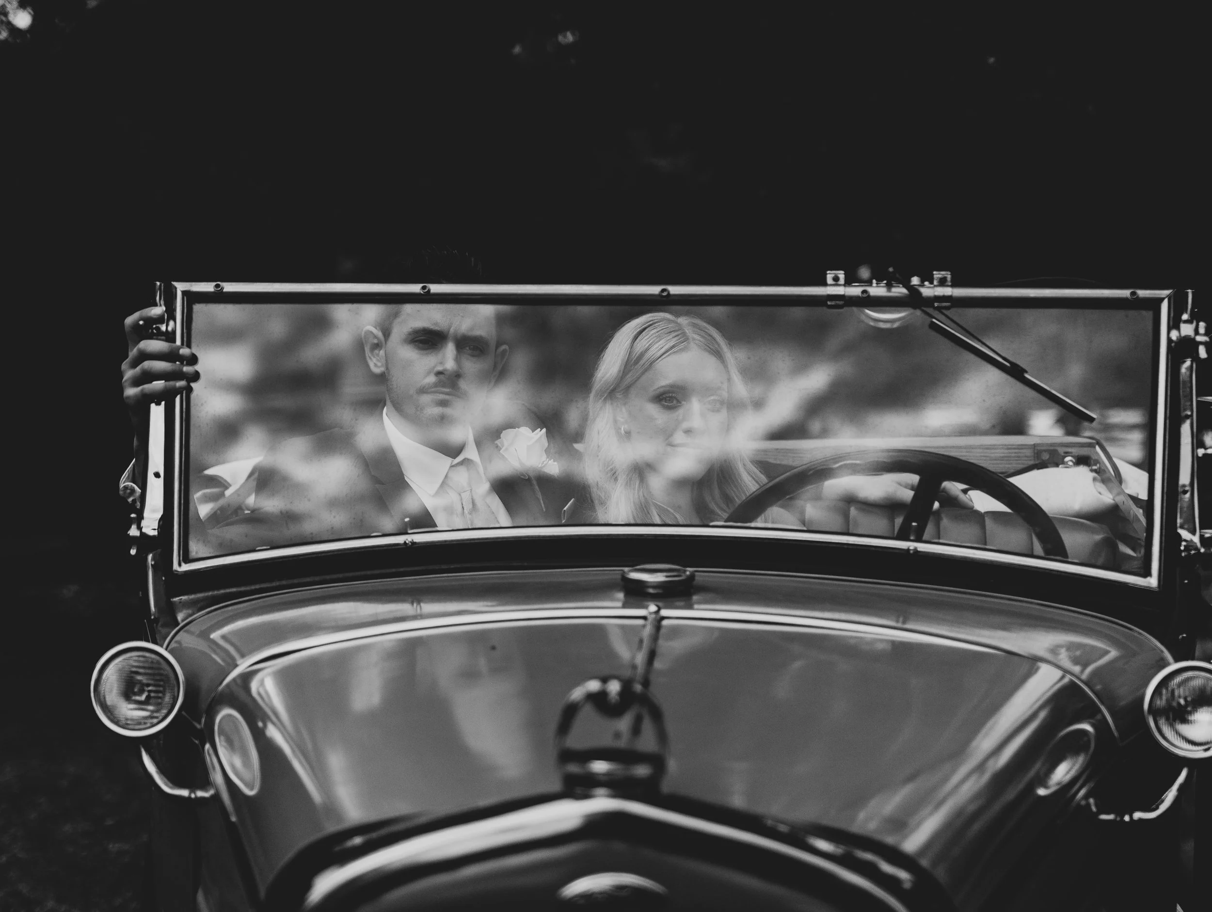 Black and white photo of a man and woman sitting in a vintage car, with the woman driving and both looking forward. The man is on the left side of the image.