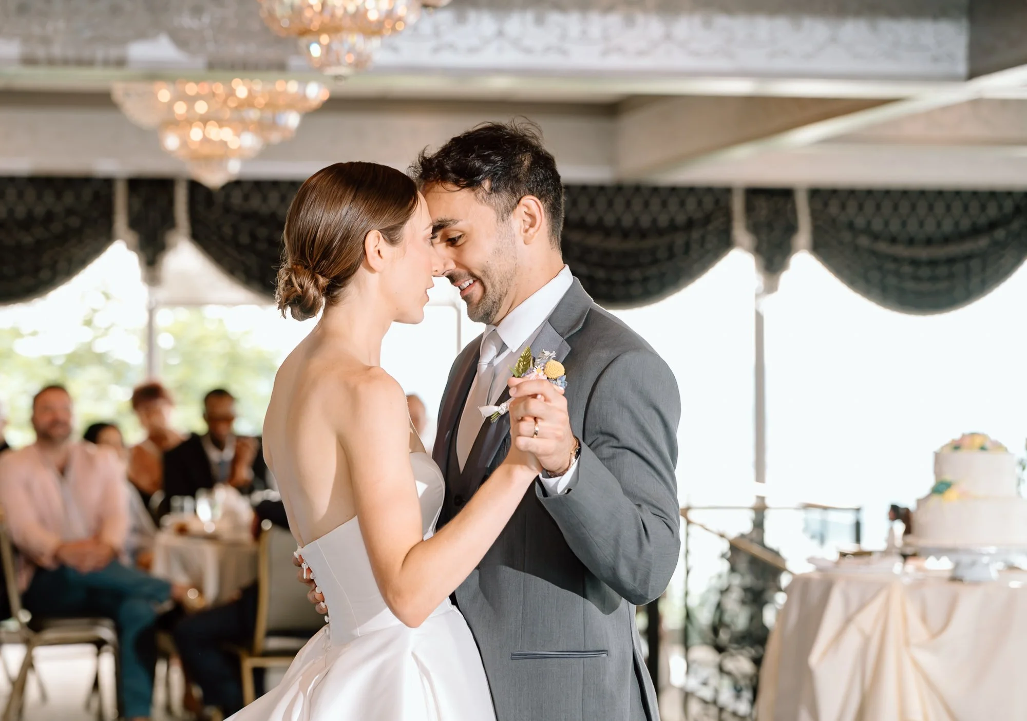 A bride and groom are sharing a dance at their wedding reception. The bride has her hair styled in a low bun and is wearing a strapless white wedding gown. The groom is dressed in a gray suit with a white shirt and tie, and is holding the bride's han