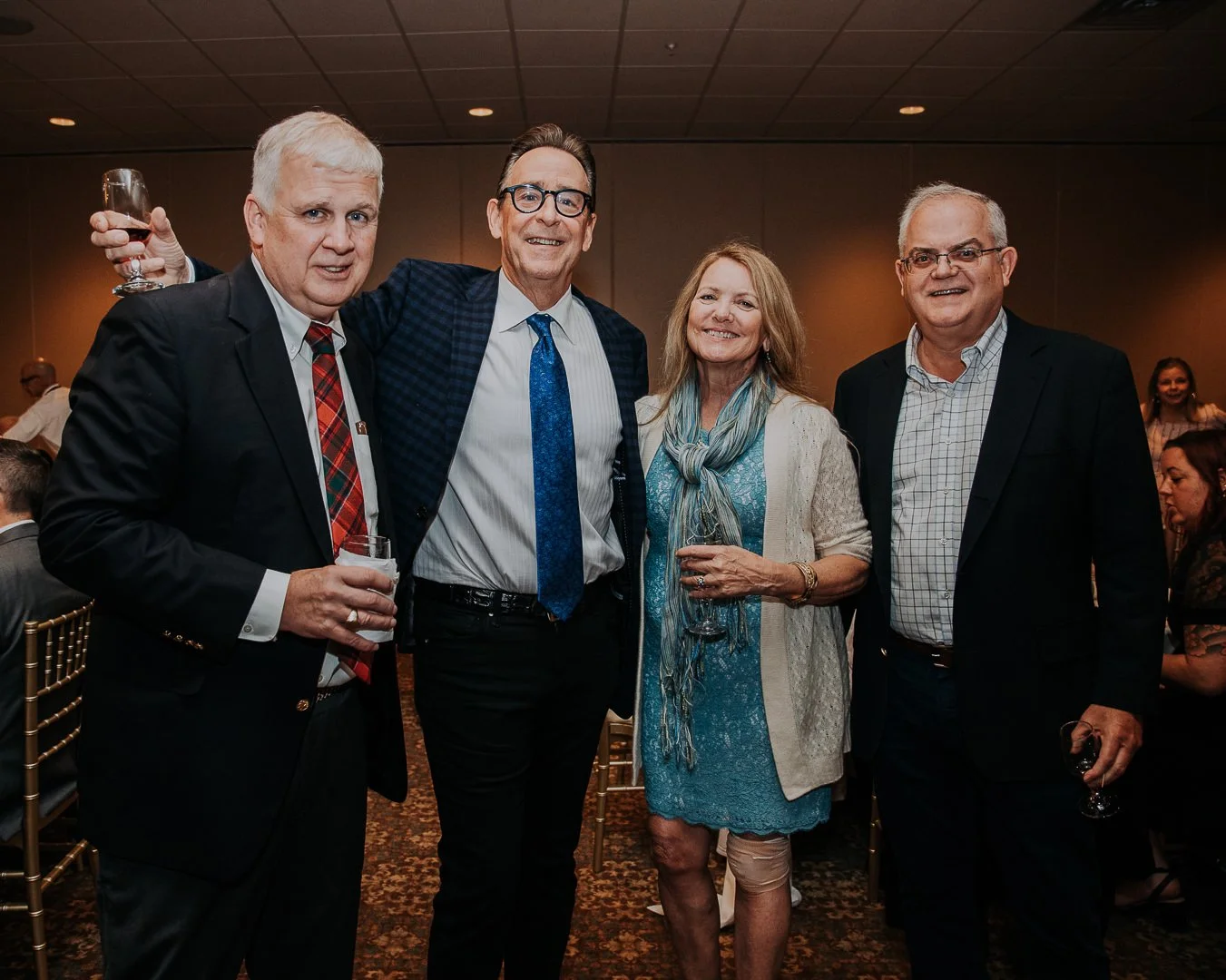 Four well-dressed people standing together at a formal event, holding glasses of wine or champagne, smiling for the camera.