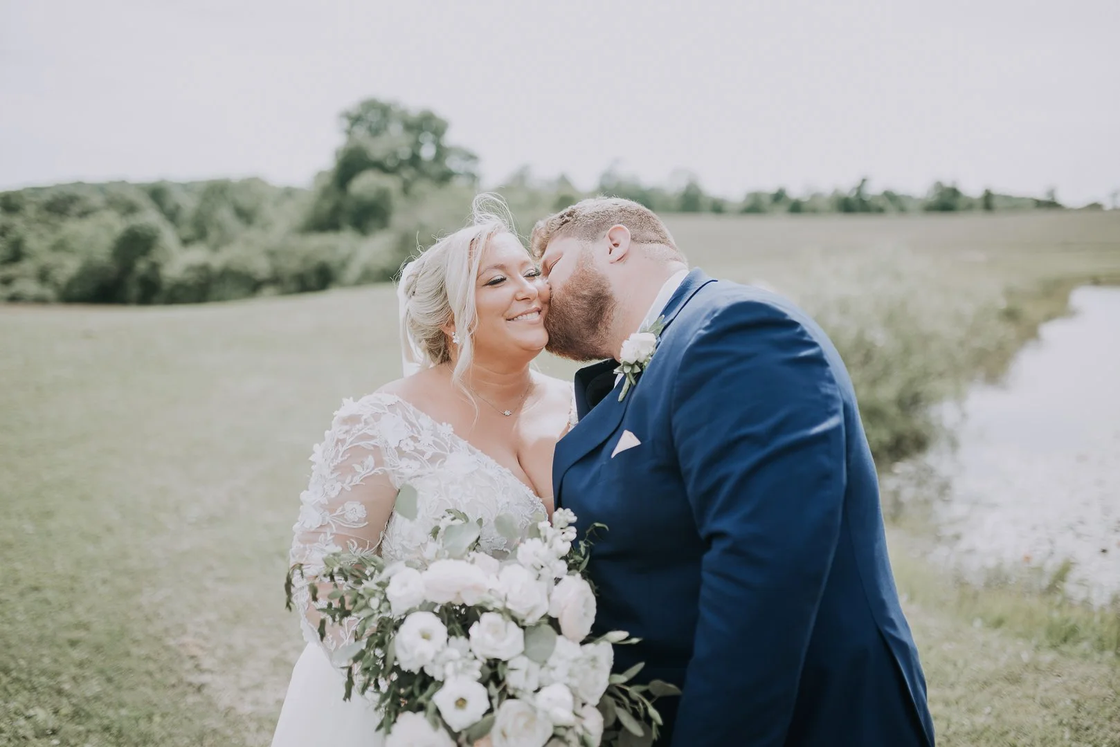 A newlywed couple standing outdoors in a grassy field, with the groom kissing the bride on the cheek. The bride is holding a bouquet of white flowers and wearing a lace wedding dress. The scene is bright and serene with a body of water and trees in t