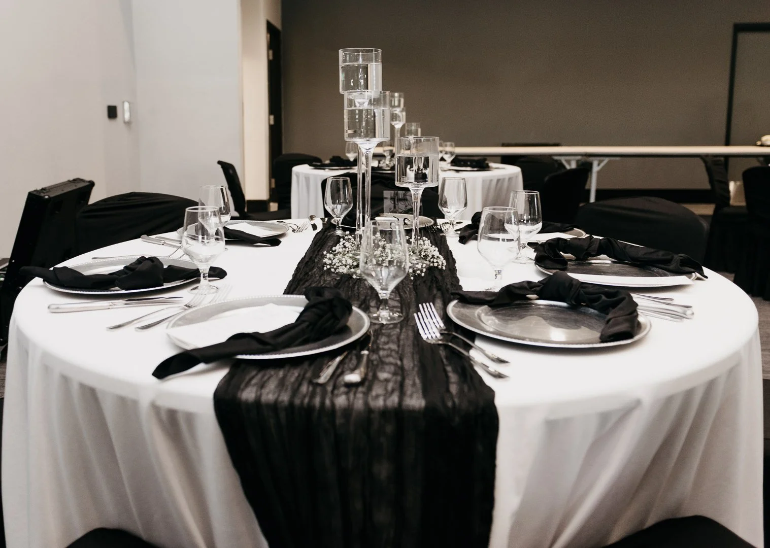 Elegant banquet table set with white tablecloth, black napkins, wine glasses, and silverware, with a black runner and tall glass centerpieces in a modern event space.