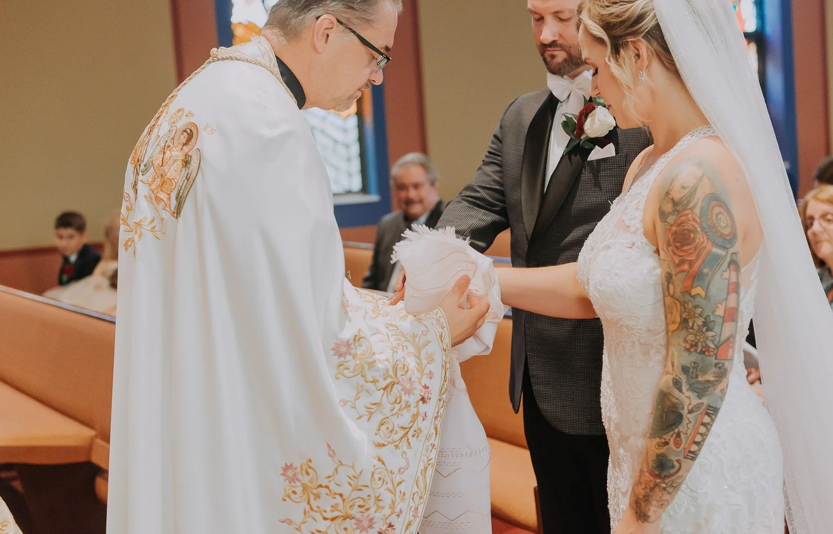 A bride and groom holding a baby during a wedding ceremony officiated by a priest inside a church. The bride has tattoos on her arm, and the priest is dressed in religious robes.