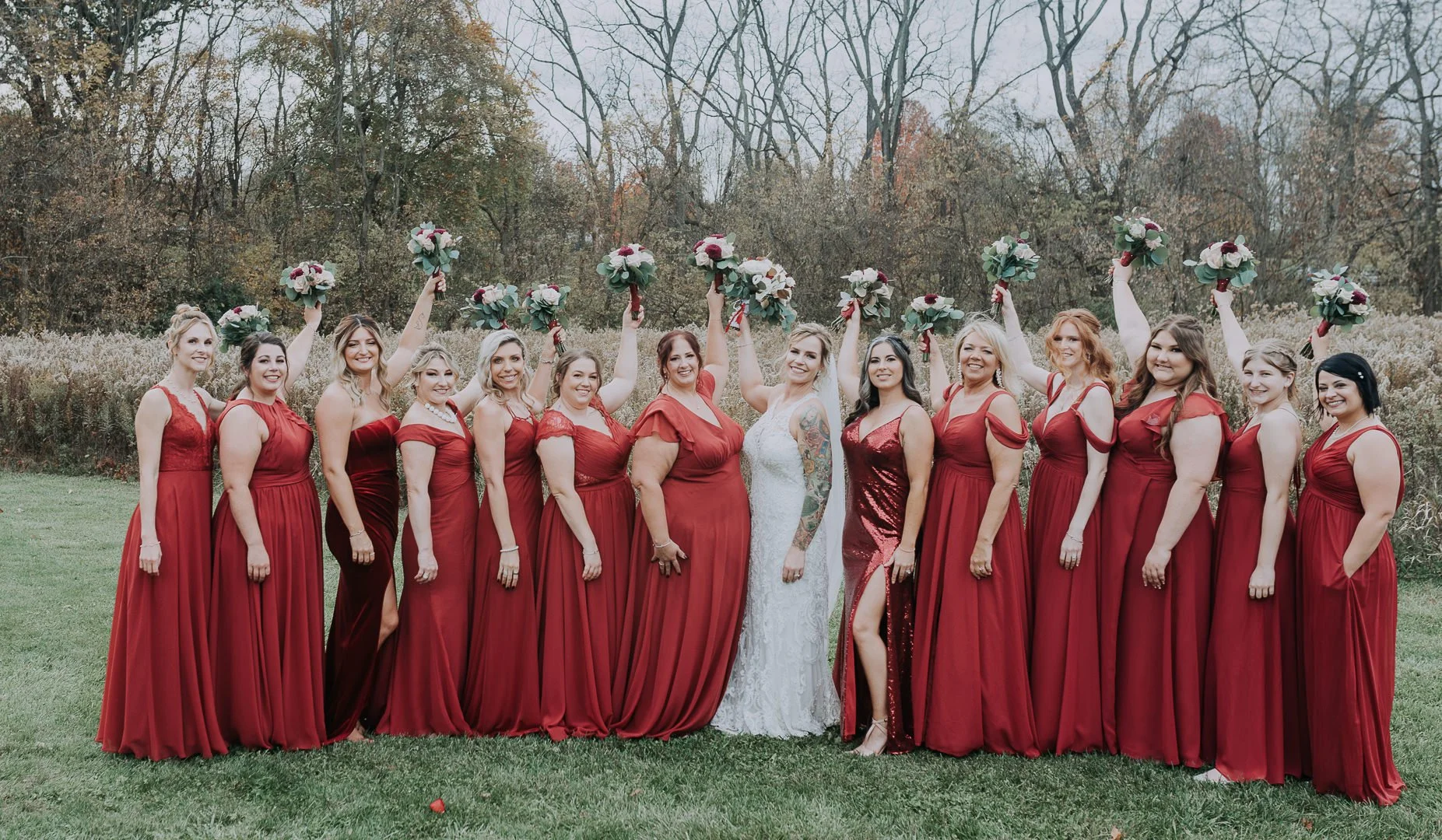 A bride and 14 bridesmaids standing outdoors on grass, all dressed in red dresses, holding bouquets of flowers, with trees in the background.