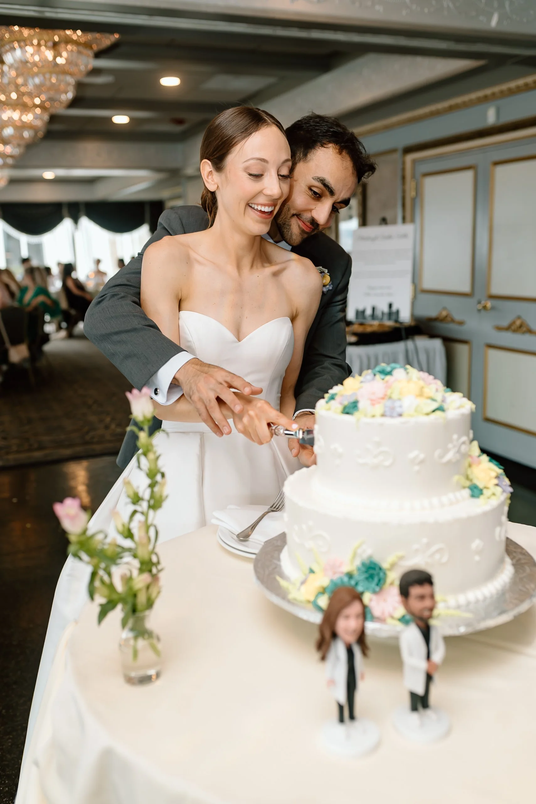 A bride and groom smiling as they cut a wedding cake, with wedding guests in the background. The cake has colorful floral decorations and some miniature bride and groom figurines on top. The table is decorated with flowers in a glass vase.