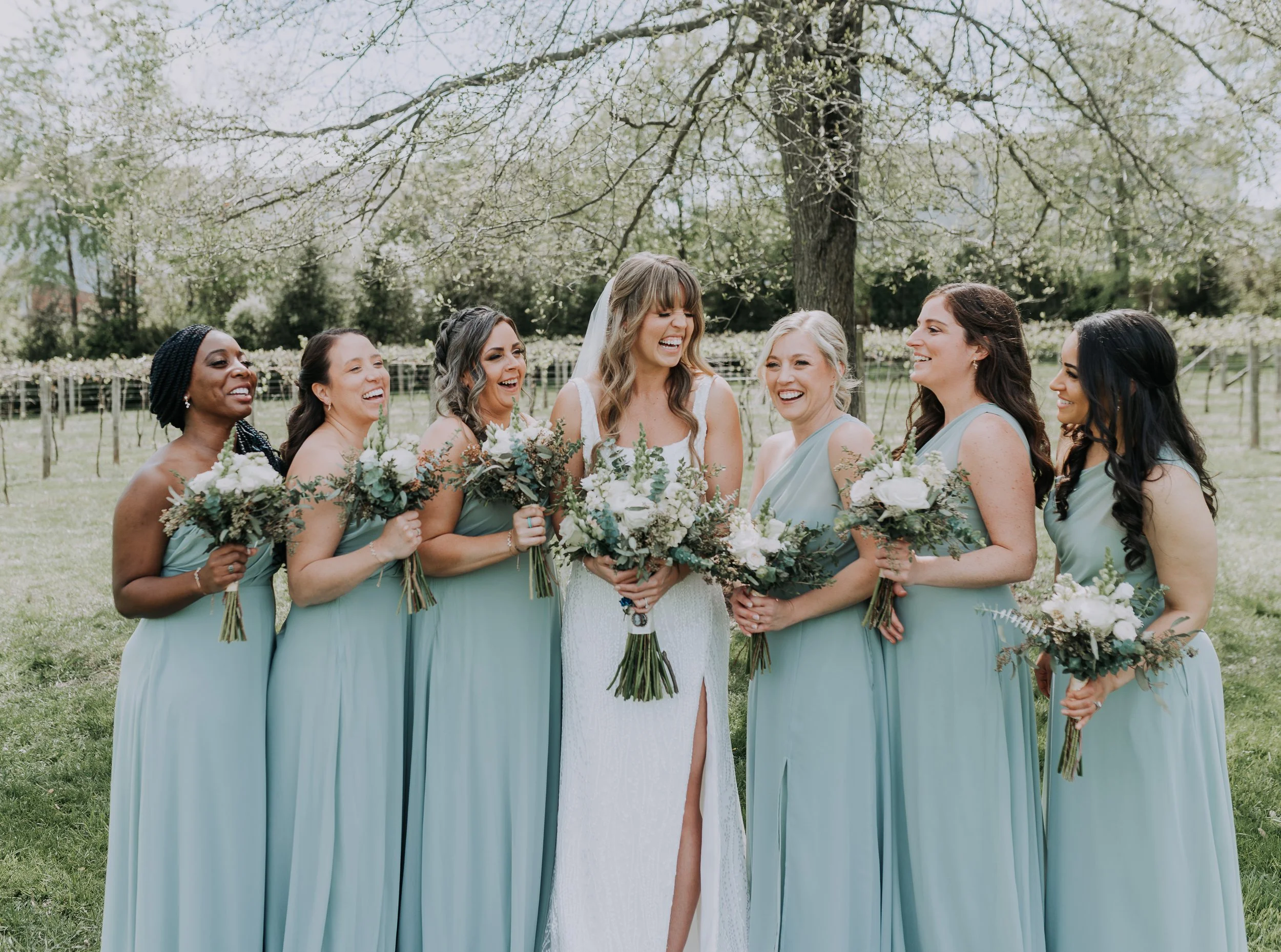 Bride and six bridesmaids standing outdoors in a park, smiling and laughing, holding bouquets of flowers, with trees and a fence in the background.