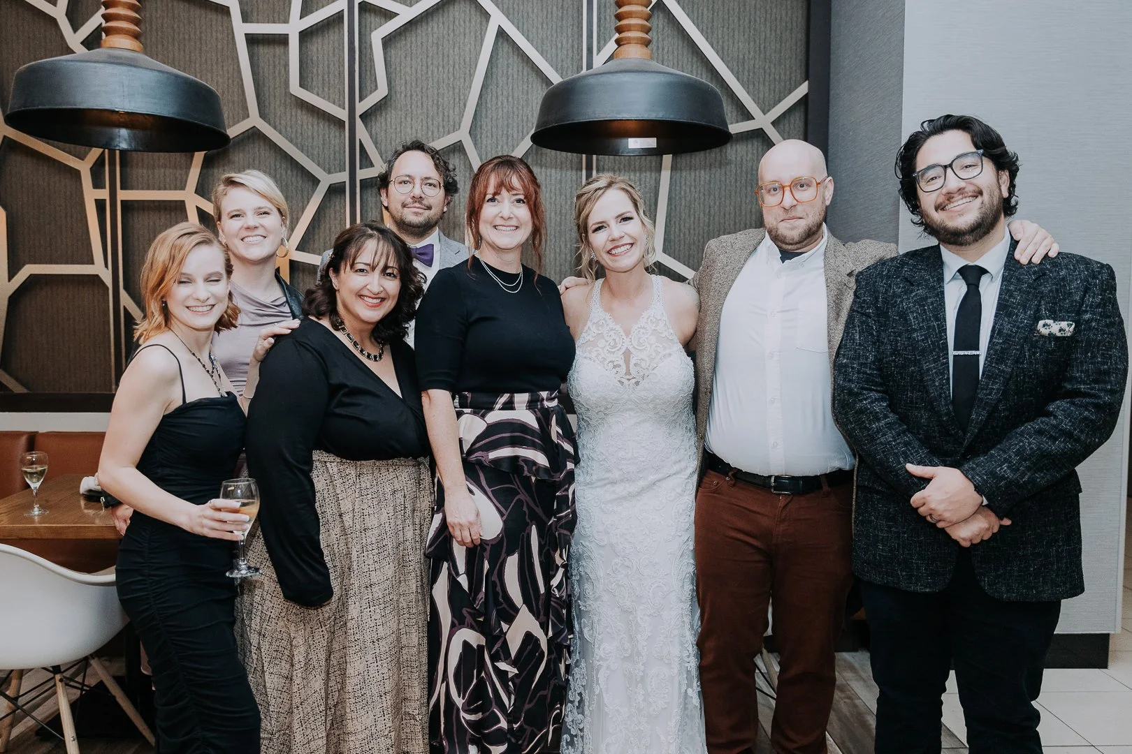 Group of nine people at a celebration, with one woman in a white lace dress in the center. They are smiling, standing close together, in a decorated room with geometric wall art and hanging black lamps.
