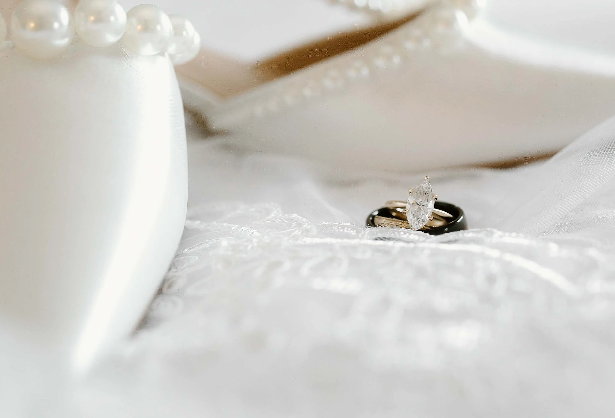 A close-up of a diamond engagement ring and a gold wedding band resting on white satin fabric, with pearls and a silver or gold object in the background.