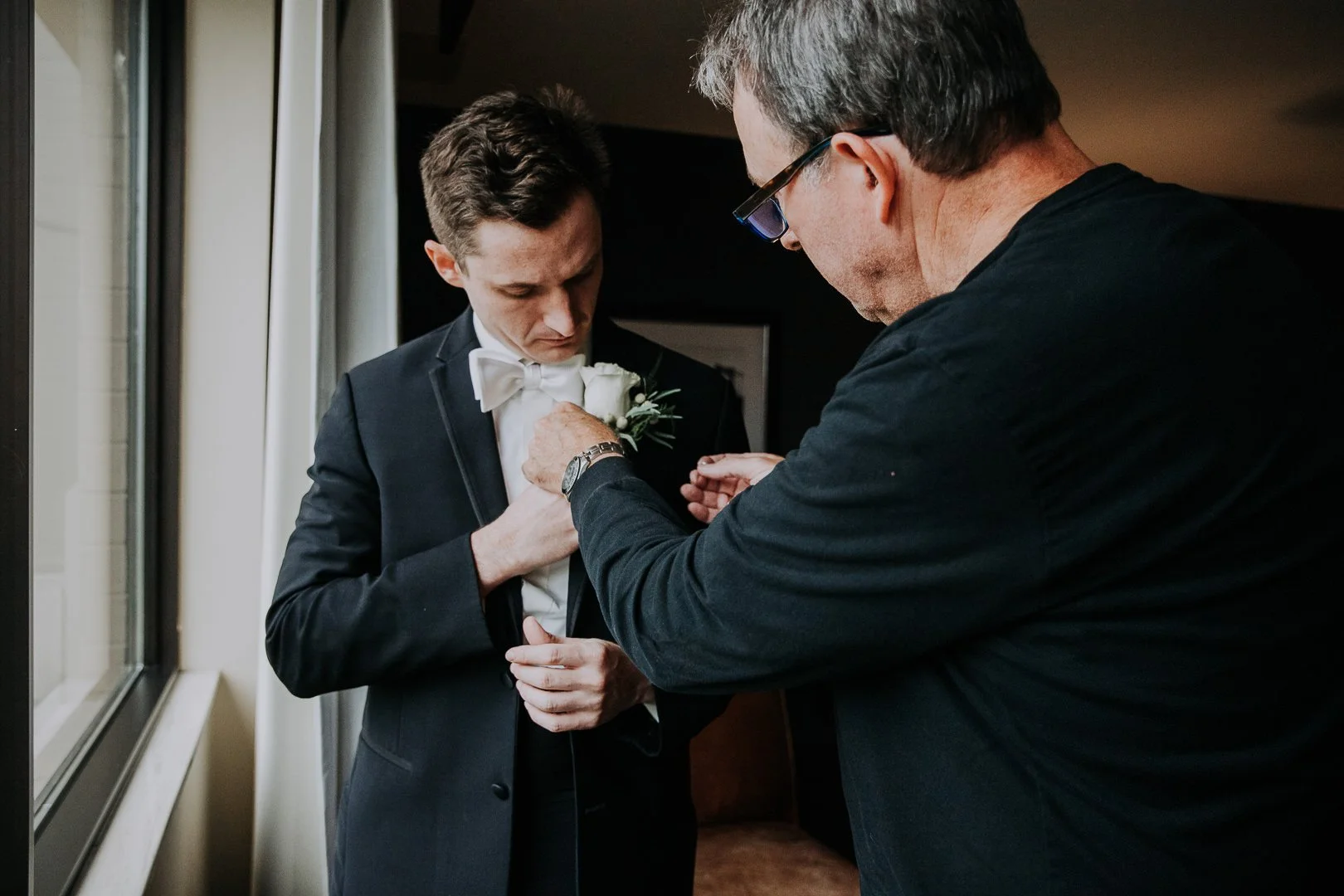 A man in a tuxedo being pinned with a boutonnière by another man.