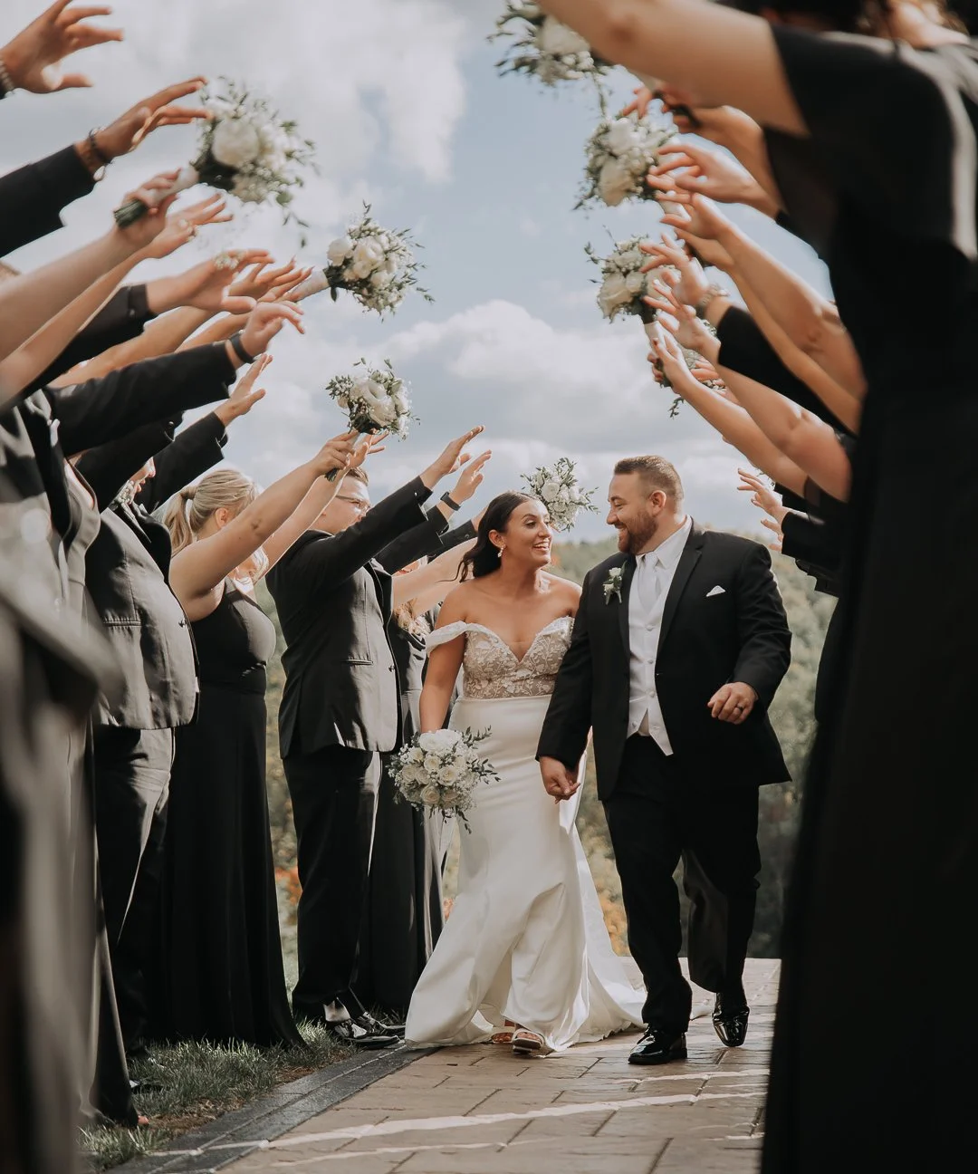 Newlywed couple walking hand in hand through a joyous crowd of bridesmaids and groomsmen holding bouquets under a cloudy sky.