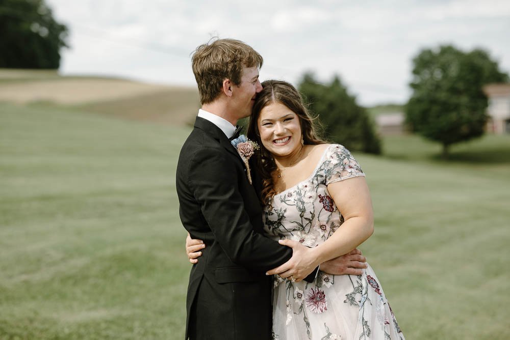 A young man and woman embrace in a green outdoor setting, smiling happily at the camera. The man is wearing a black suit, while the woman is in a light-colored dress with floral patterns. The background features grassy hills and trees under a partly