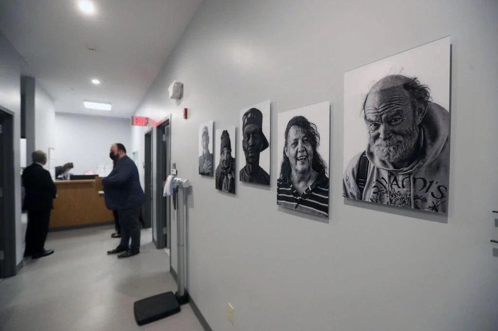 The image shows a hallway in a building, with artwork featuring black-and-white portraits of diverse people on the wall. At the end of the hall, there is a reception desk where two individuals are engaged in conversation. The scene appears to be insi