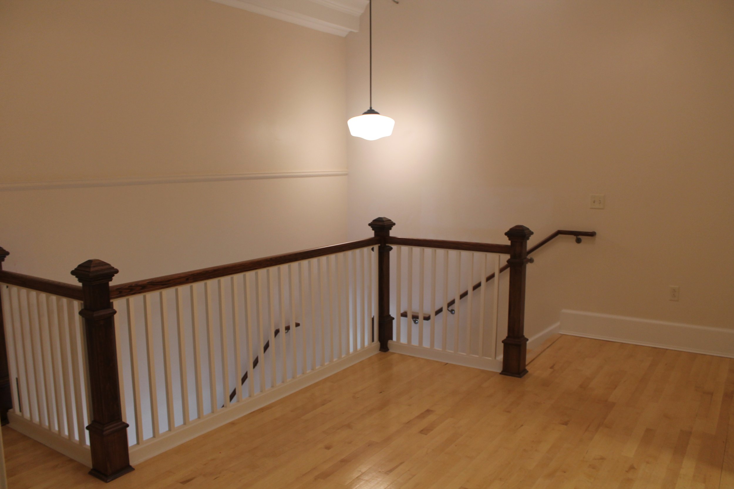 Empty staircase landing with wooden handrails, white spindles, wood floor, and a hanging ceiling light.