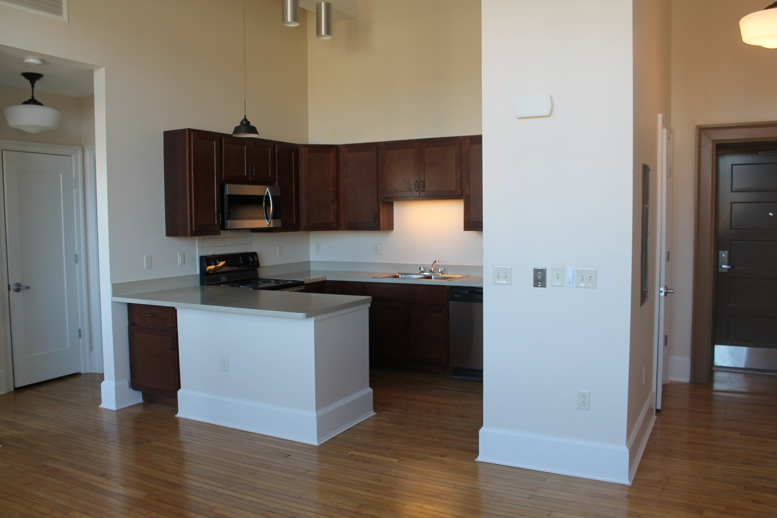 Empty kitchen with dark brown cabinets, a white countertop, and hardwood flooring, with a doorway leading to a darker hallway.