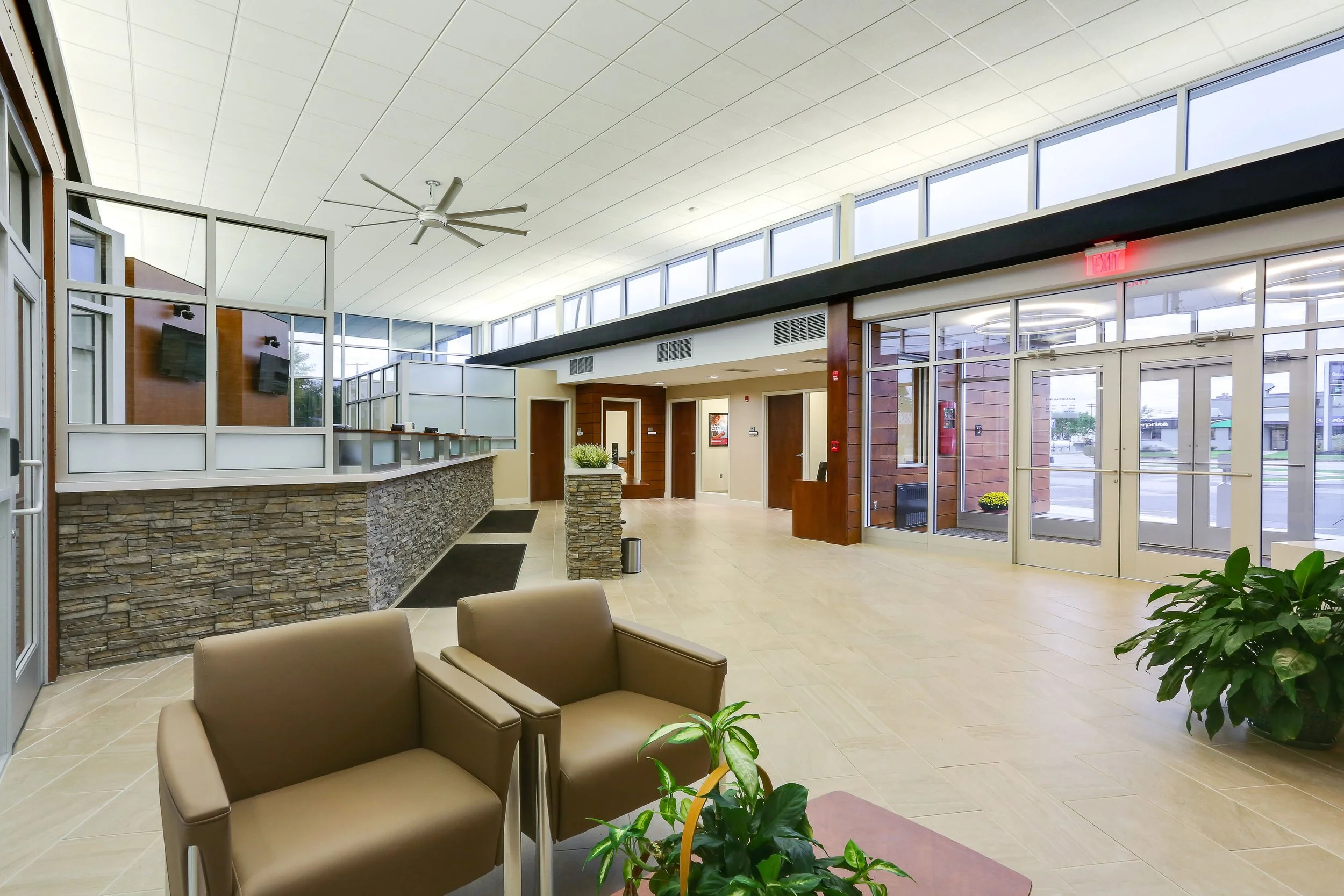 Modern hotel lobby with beige chairs, potted plants, large front window, and check-in desk with stone facade.