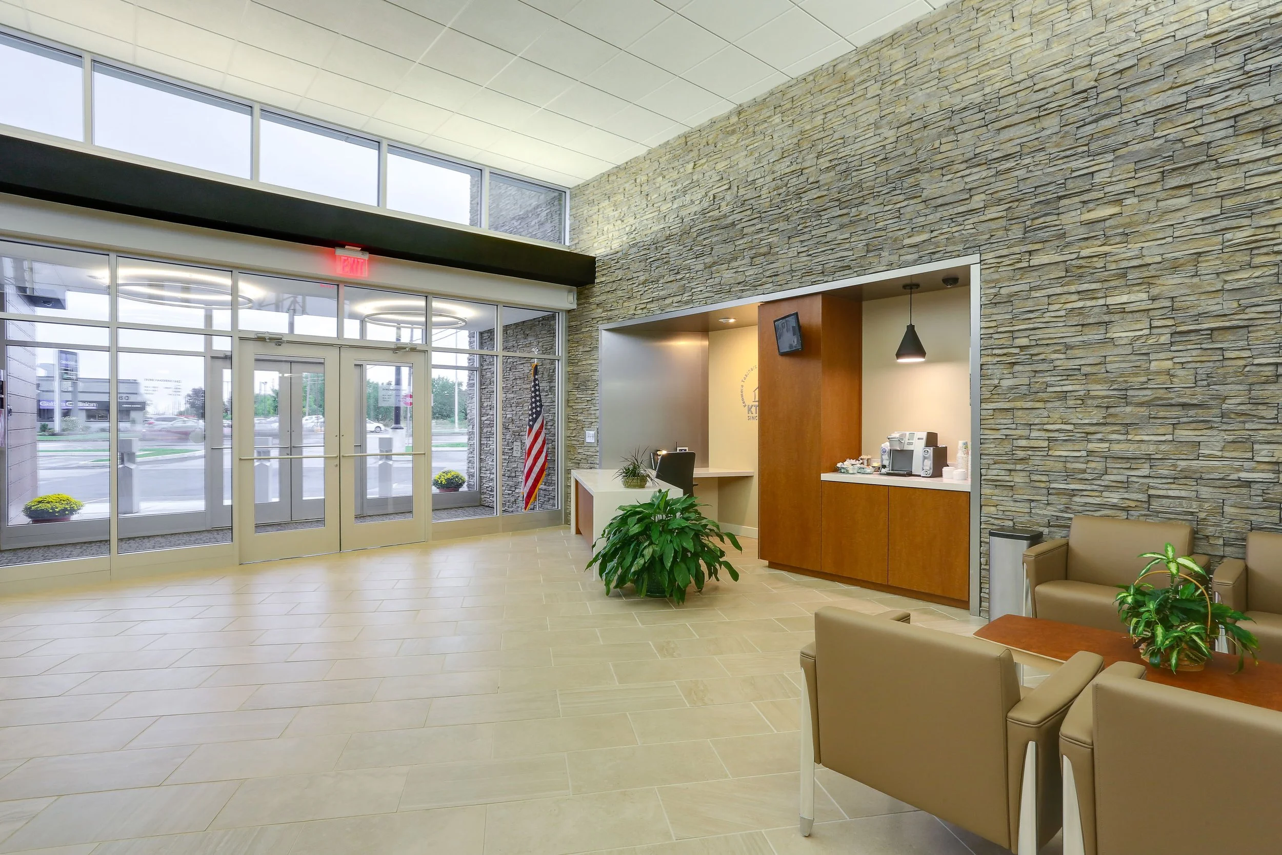 Interior of a modern building lobby with glass entrance doors, beige tiled floor, stone accent wall, reception desk, coffee station, and seating area with beige chairs and green plants.