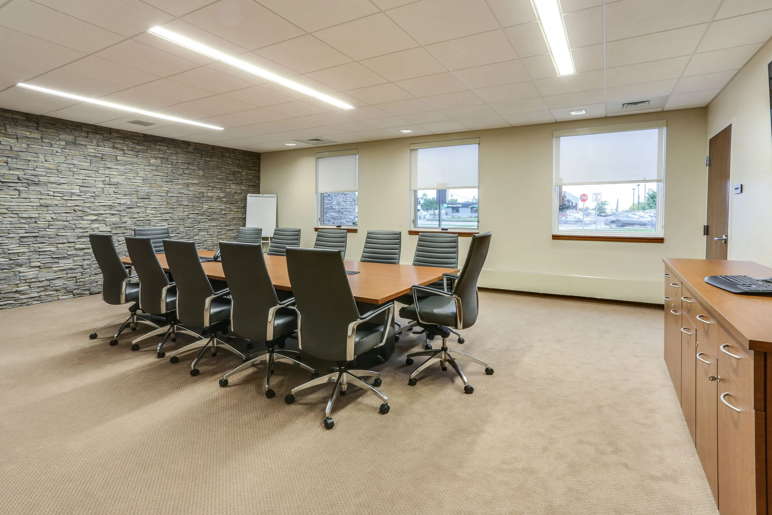 Empty conference room with a large wooden table surrounded by black ergonomic chairs, stone accent wall, beige carpet, window blinds, and a cabinet with a computer keyboard.