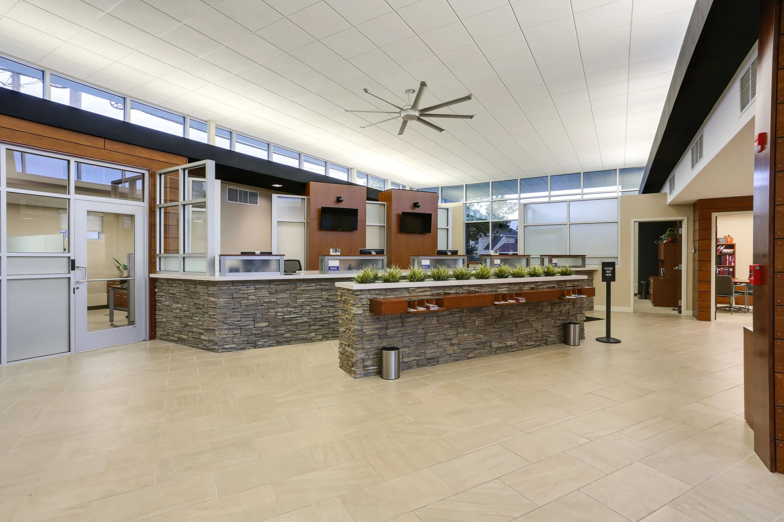 Empty hotel or office reception area with a front desk, plant decorations, multiple computer stations, three wall-mounted TVs, and seating areas in the background.