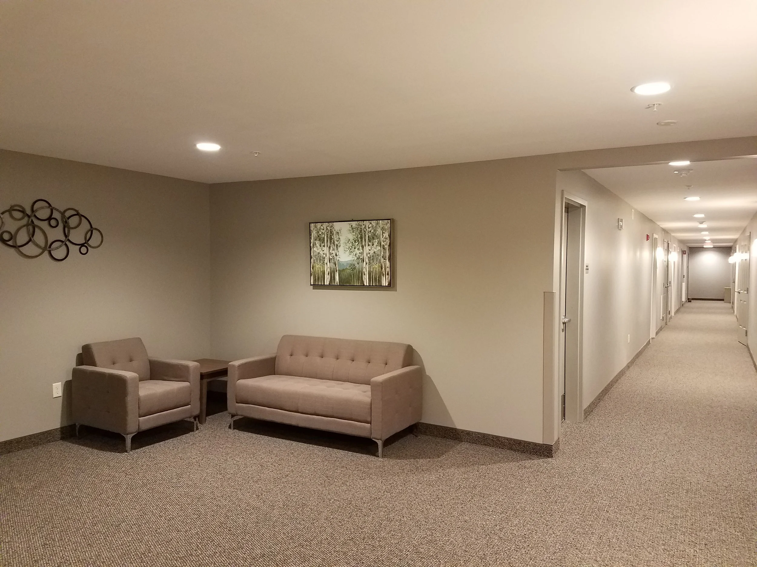Empty hotel lobby or waiting area with two beige armchairs, a matching beige sofa, a small side table, wall art, and modern lighting, with a hallway in the background.