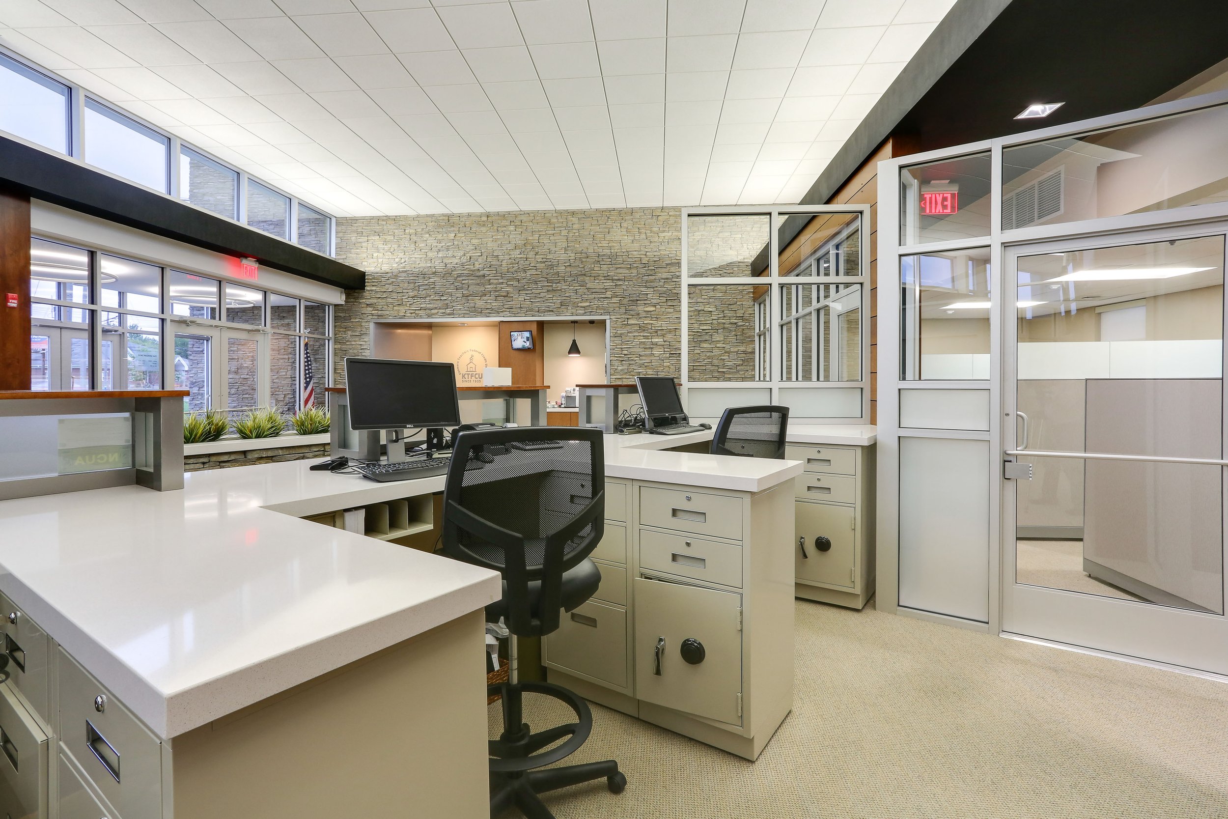 Modern office reception area with desks, computers, office chairs, and a stone accent wall with an entrance door and large windows.