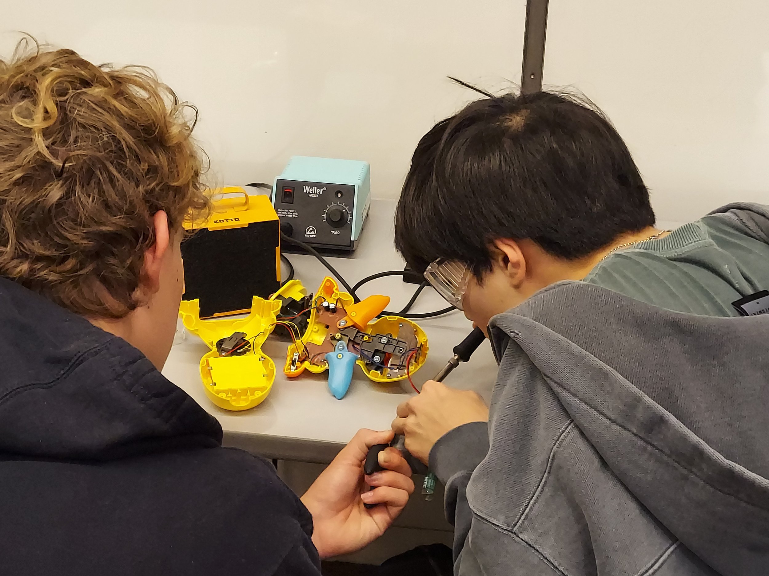 Two students at a worktable soldering an accessible switch to a toy.