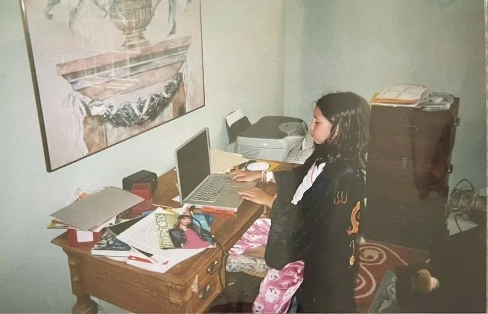A young girl standing at a cluttered wooden desk with a laptop, papers, magazines, and office supplies, in a room with a painting on the wall and a small filing cabinet.