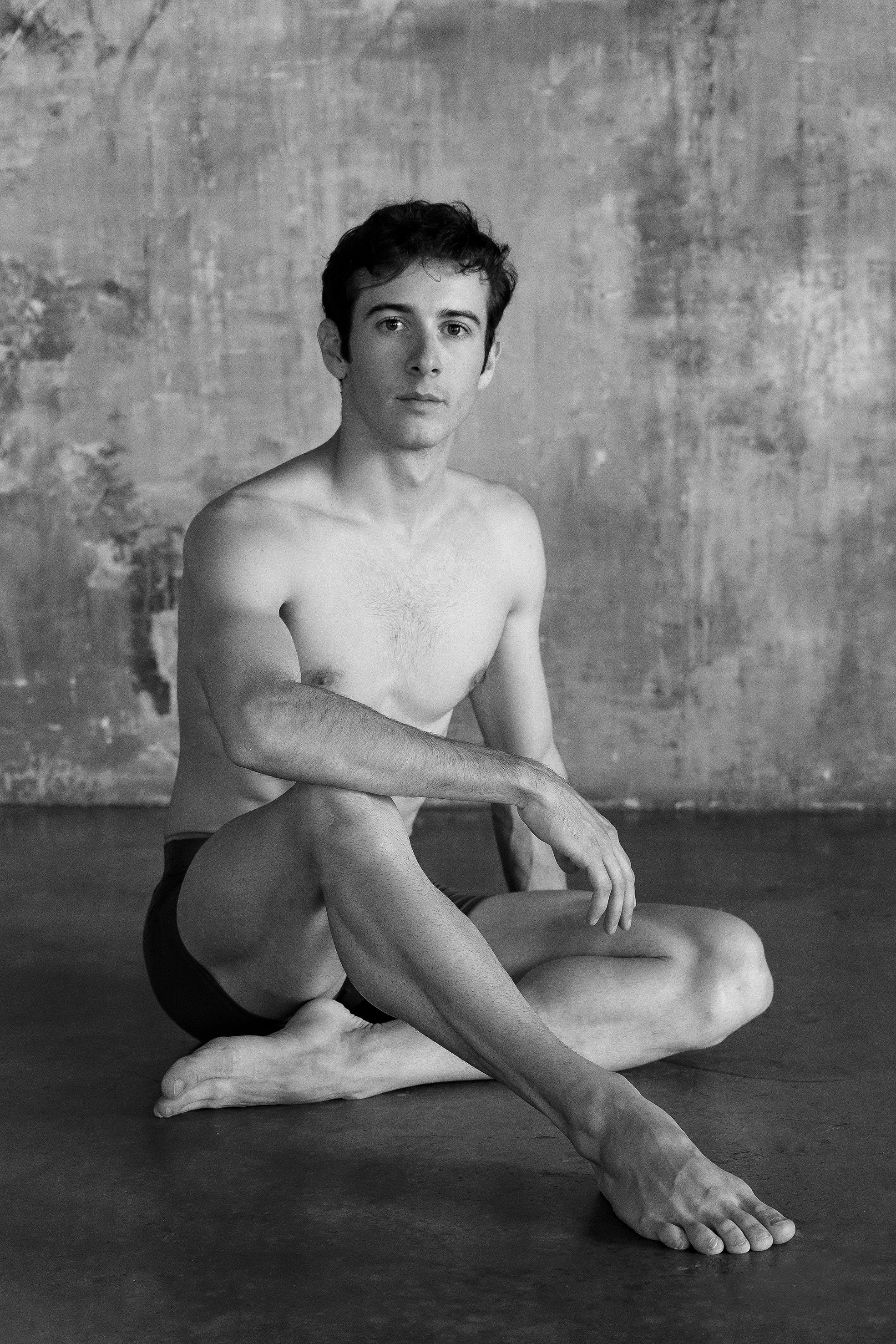 A black-and-white photo of two young men with lean physiques, sitting on a dark floor against a textured wall. One man is seated with his legs crossed, the other is kneeling, and the man in the cross-legged position is resting his arm on his knee, looking directly at the camera with a neutral expression.