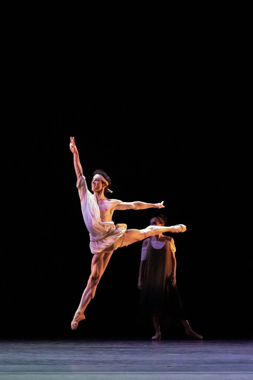 A male ballet dancer in light-colored costume performing a leap on stage with a woman in dark clothing standing in the background.