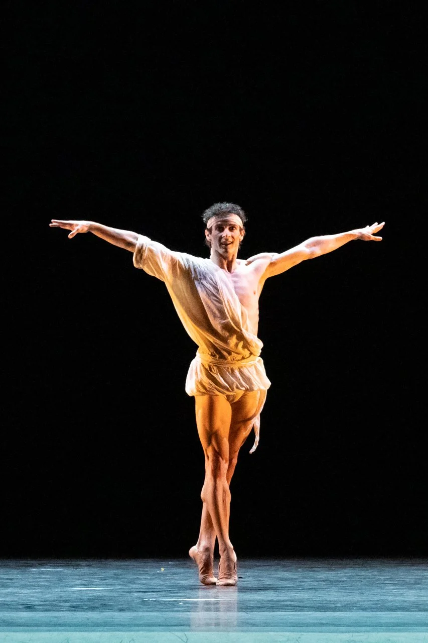 A male ballet dancer posing on stage with arms extended, wearing a loose beige costume, against a black background.