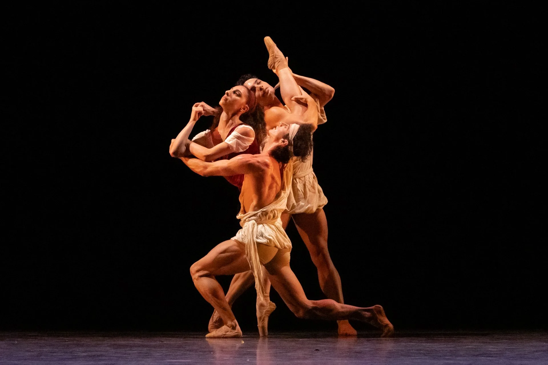 Four ballet dancers in earthy-toned costumes perform on a dark stage in an intricate pose.