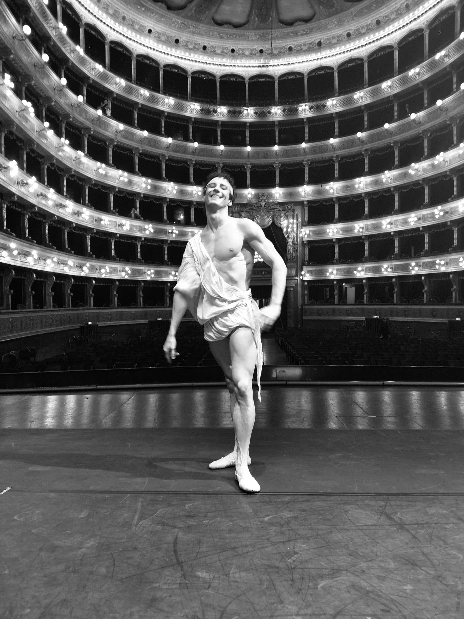 Black and white photo of a ballet dancer on stage in a theater with multiple balconies and lights, smiling with arms in a ballet pose.