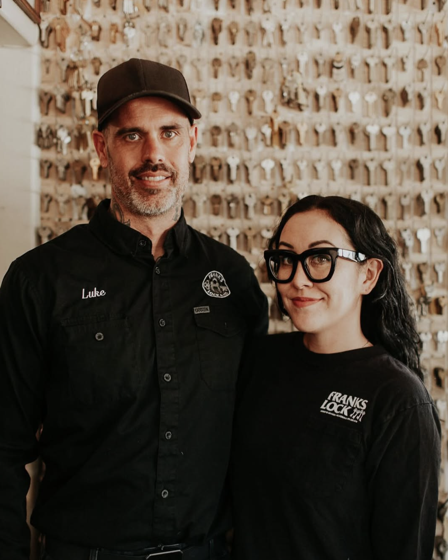 A man and a woman standing together in front of a wall filled with keys. The man is wearing a black shirt with a name tag that says 'Luke' and a logo, and a cap. The woman has black glasses, curly black hair, and a black shirt with a logo that says '