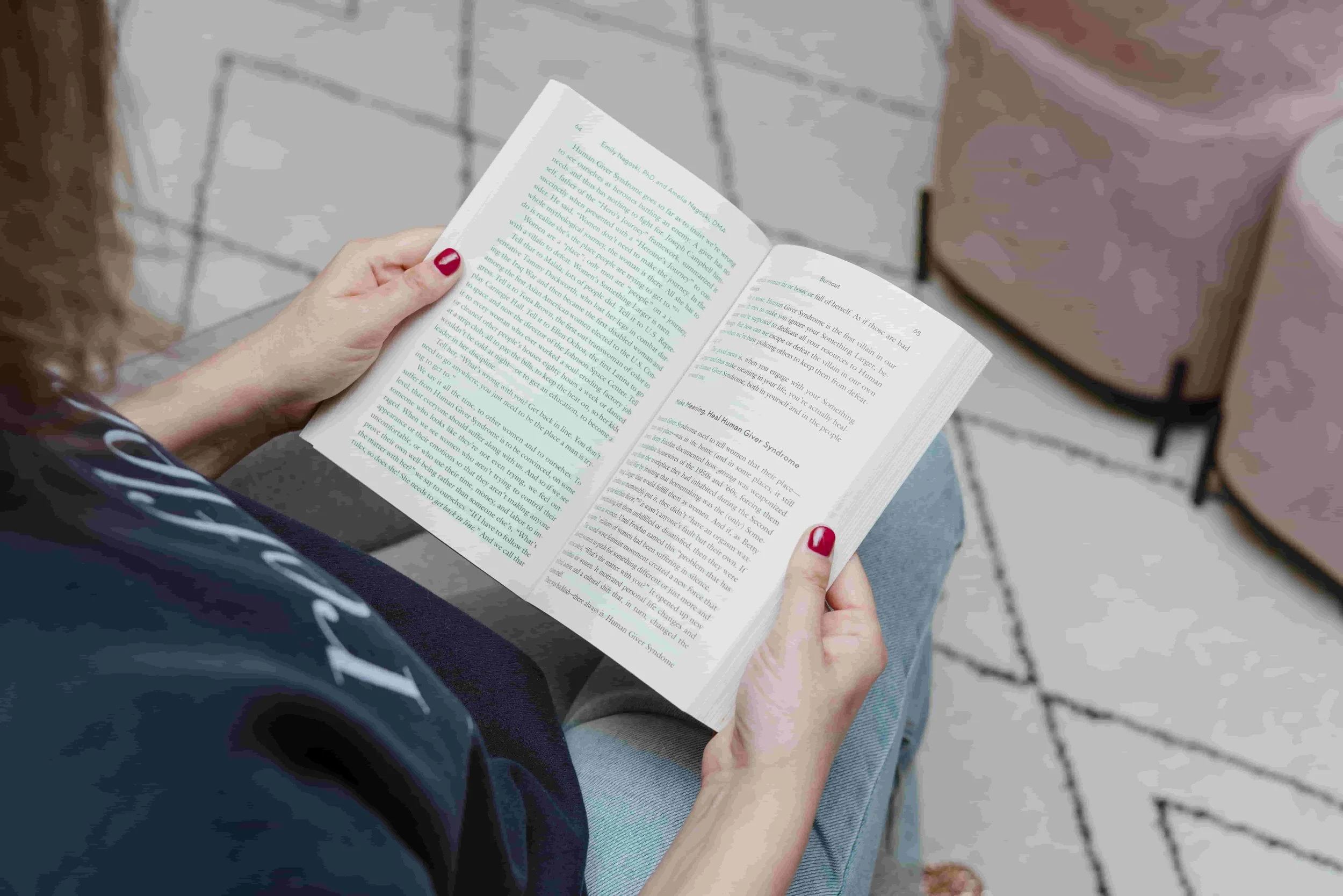 A person with red nail polish reading a book while seated, with pink upholstered chairs and a patterned carpet in the background.