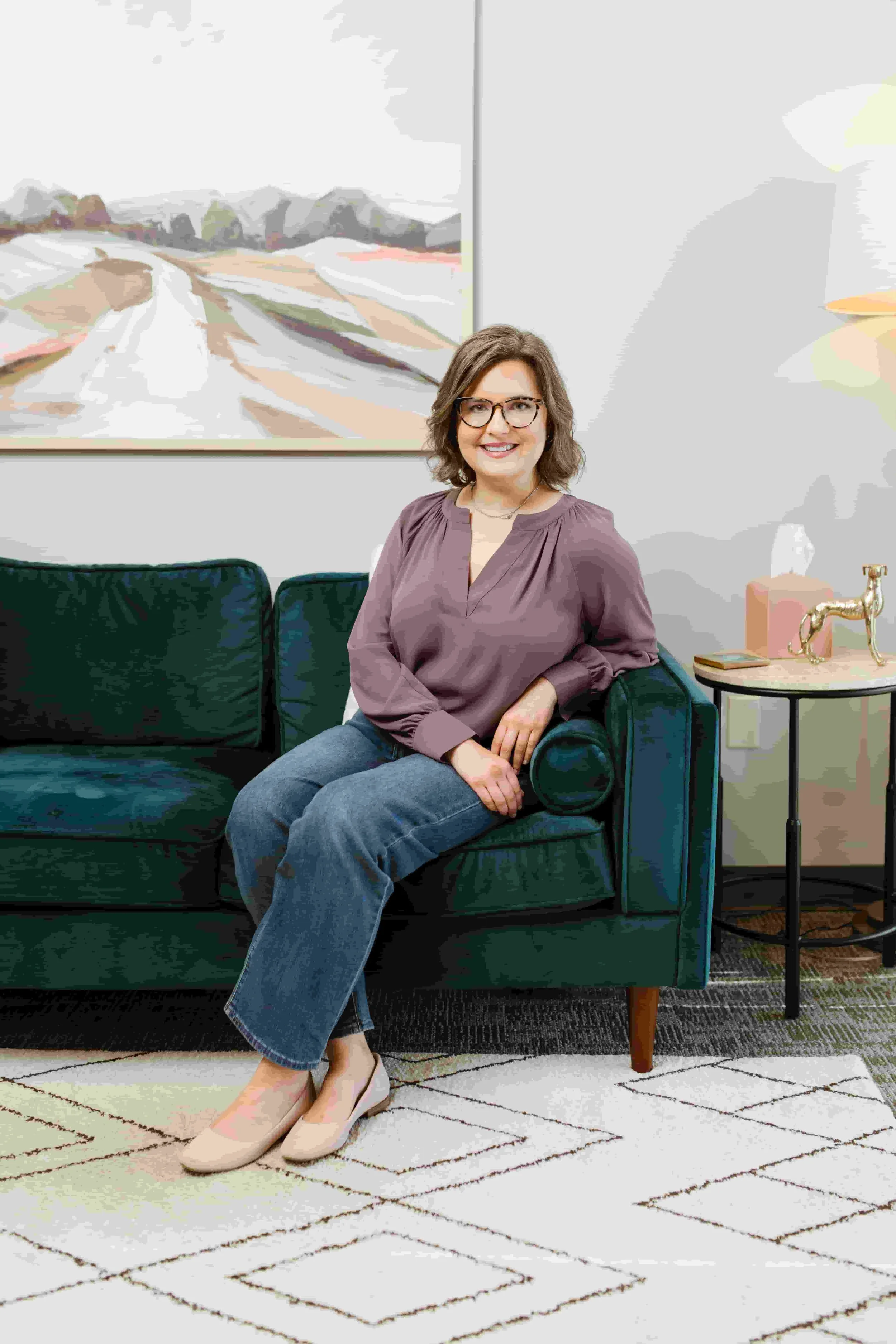 A woman with shoulder-length brown hair, wearing glasses, a mauve blouse, and blue jeans, sitting indoors with a smile.