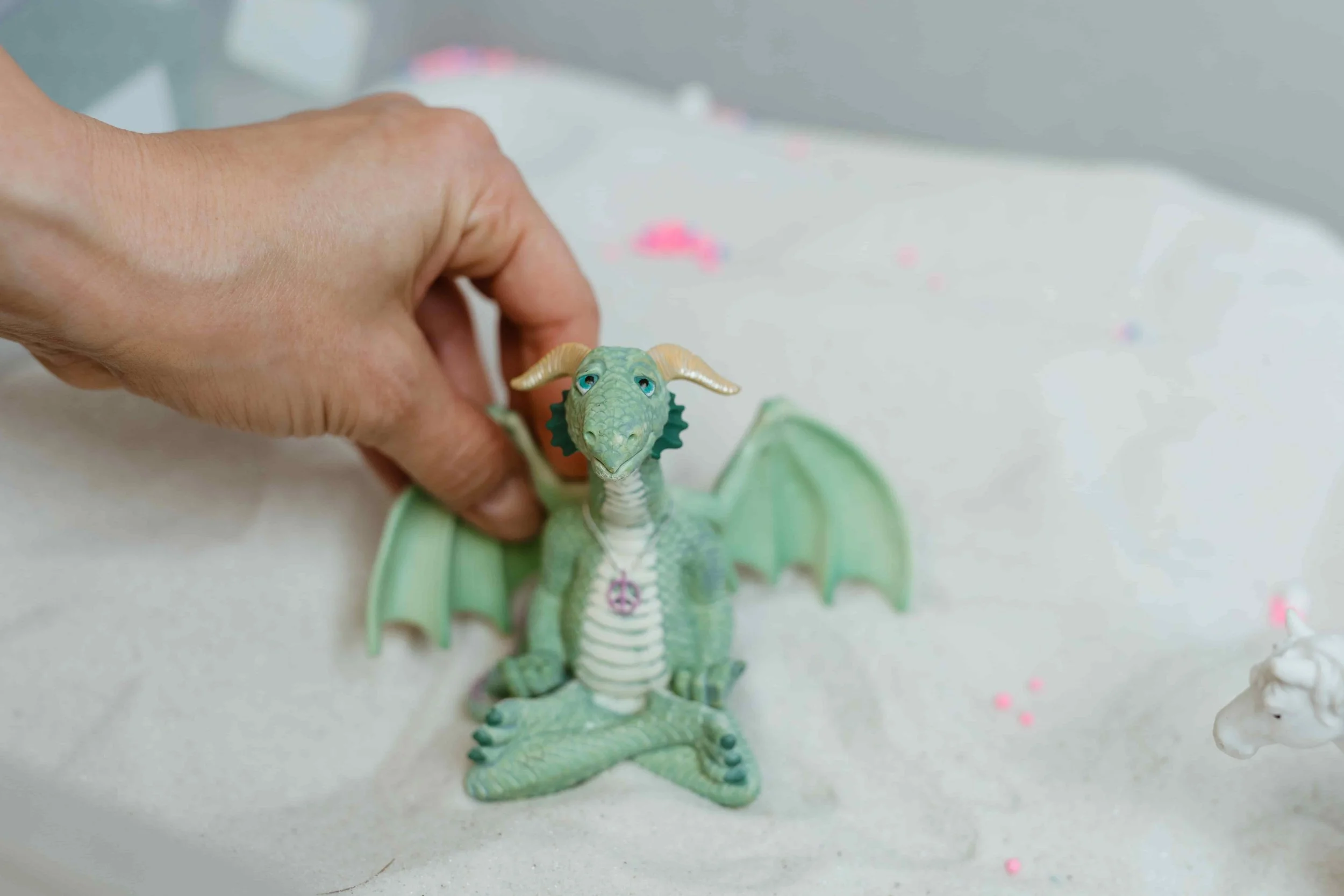 A person’s hand holding a green dragon figurine with wings, horns, and a necklace, sitting on white sand with small pink and white decorations, and a small white unicorn figurine in the background.