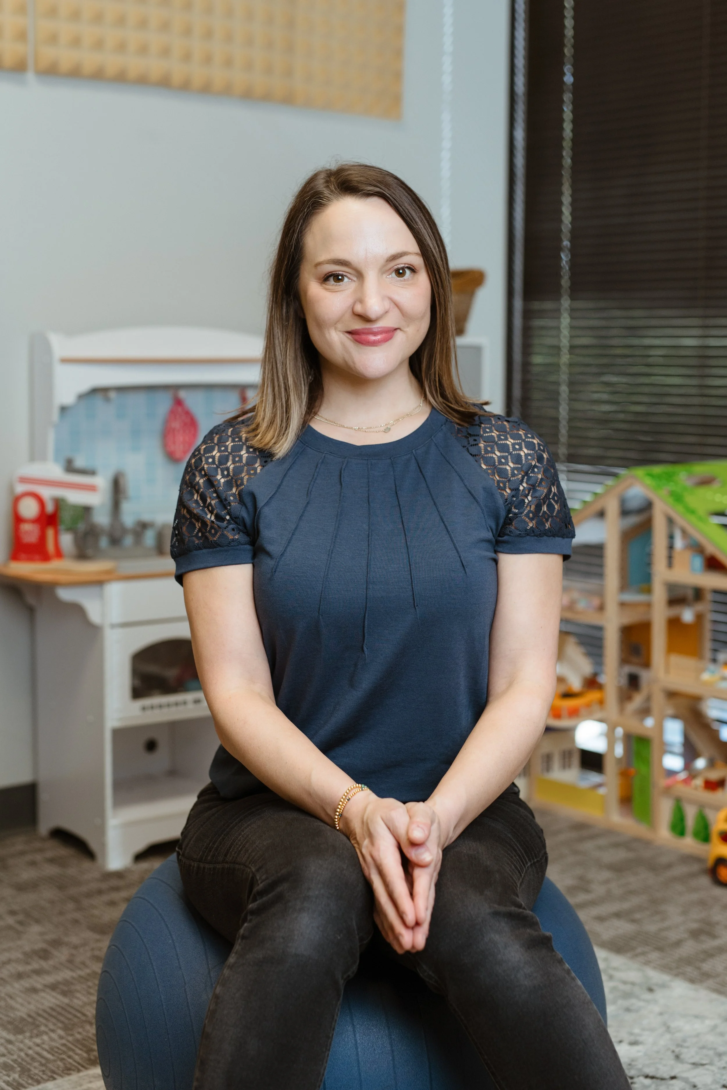 A smiling woman with shoulder-length brown hair wearing a navy blue top with lace and pleats, standing indoors against an abstract blue and gray background.