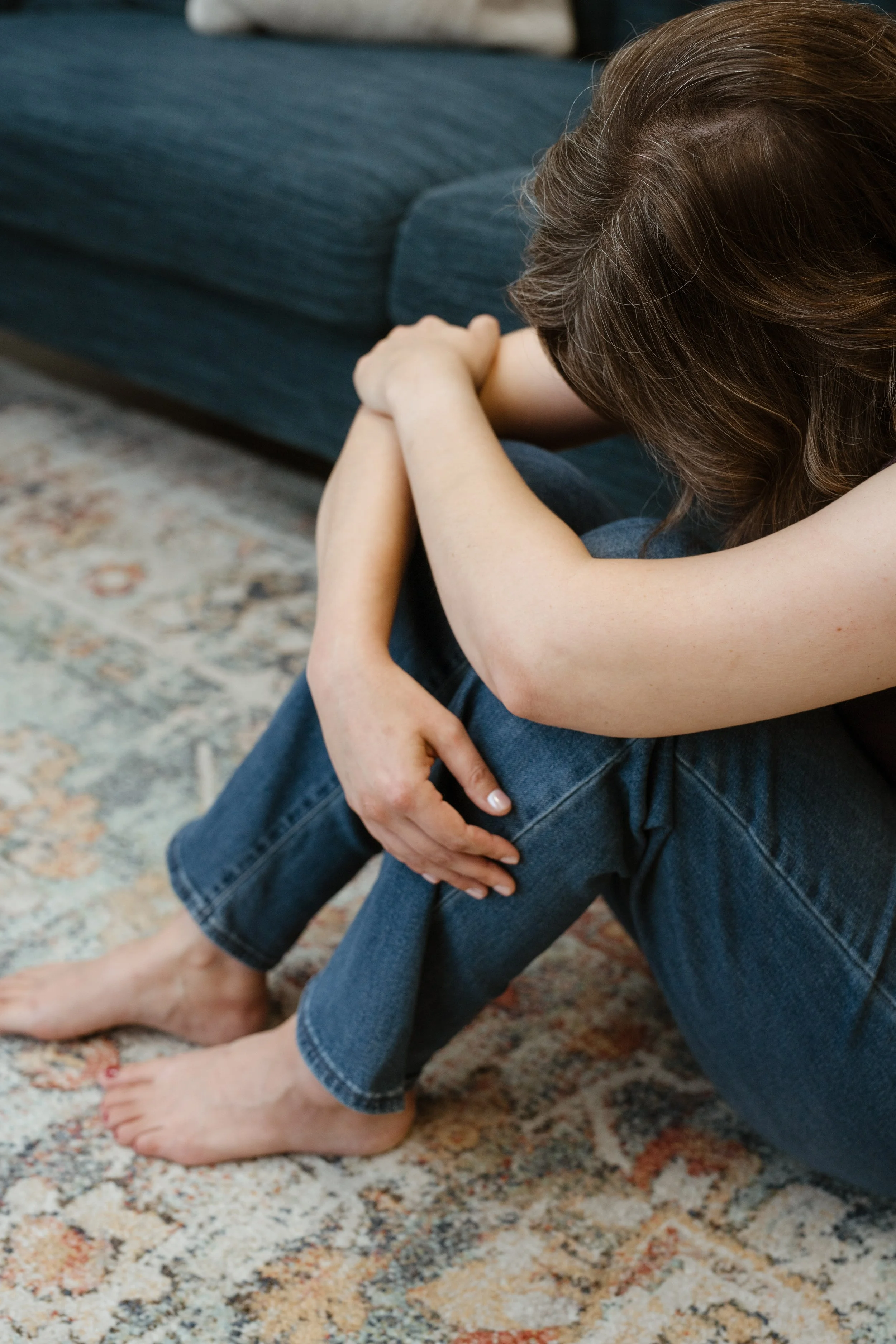 A woman sitting on the floor with her head down and arms crossed over her knees in a sign of distress or sadness, in a living room setting.