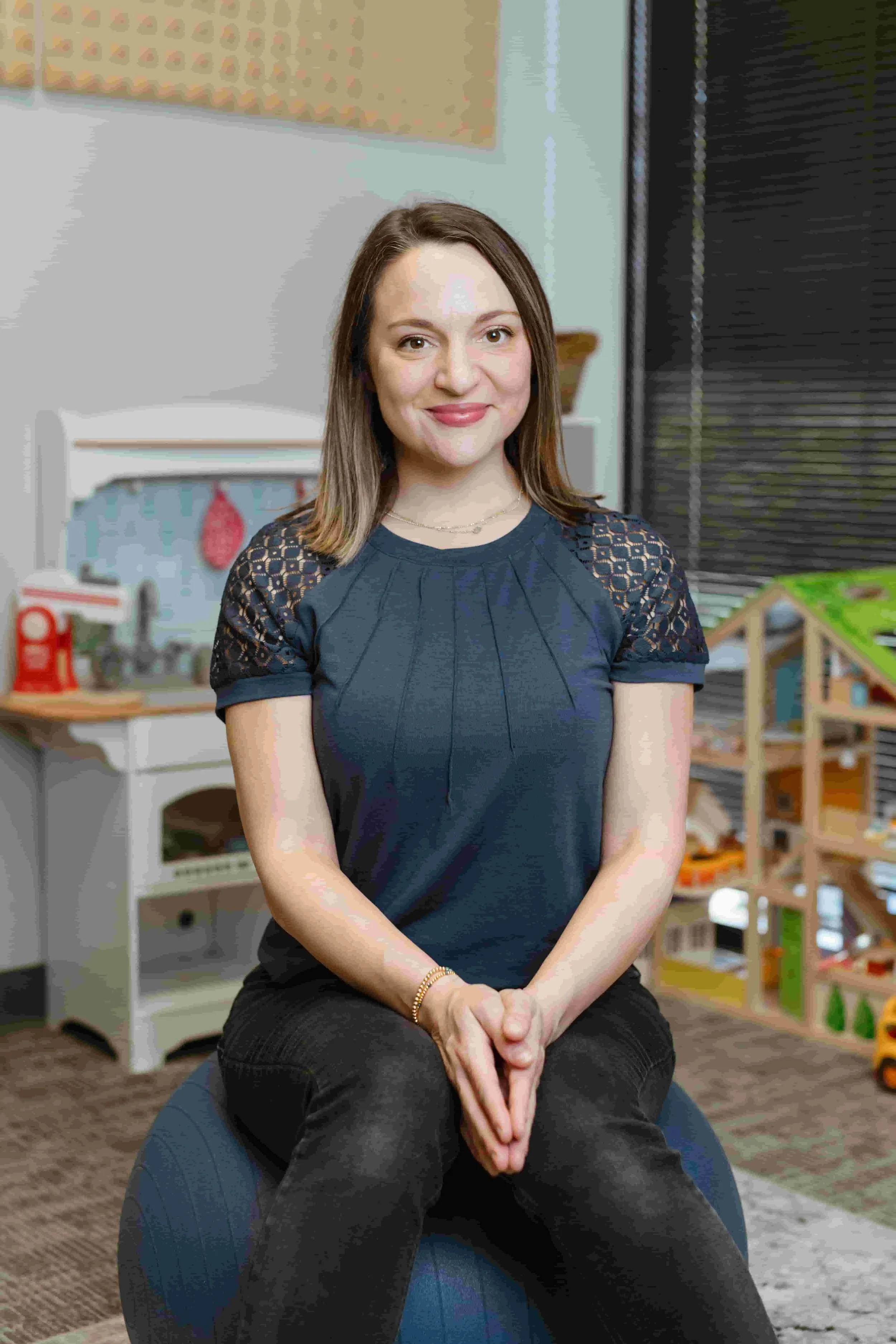 A smiling woman with shoulder-length brown hair wearing a navy blue top with lace and pleats, standing indoors against an abstract blue and gray background.