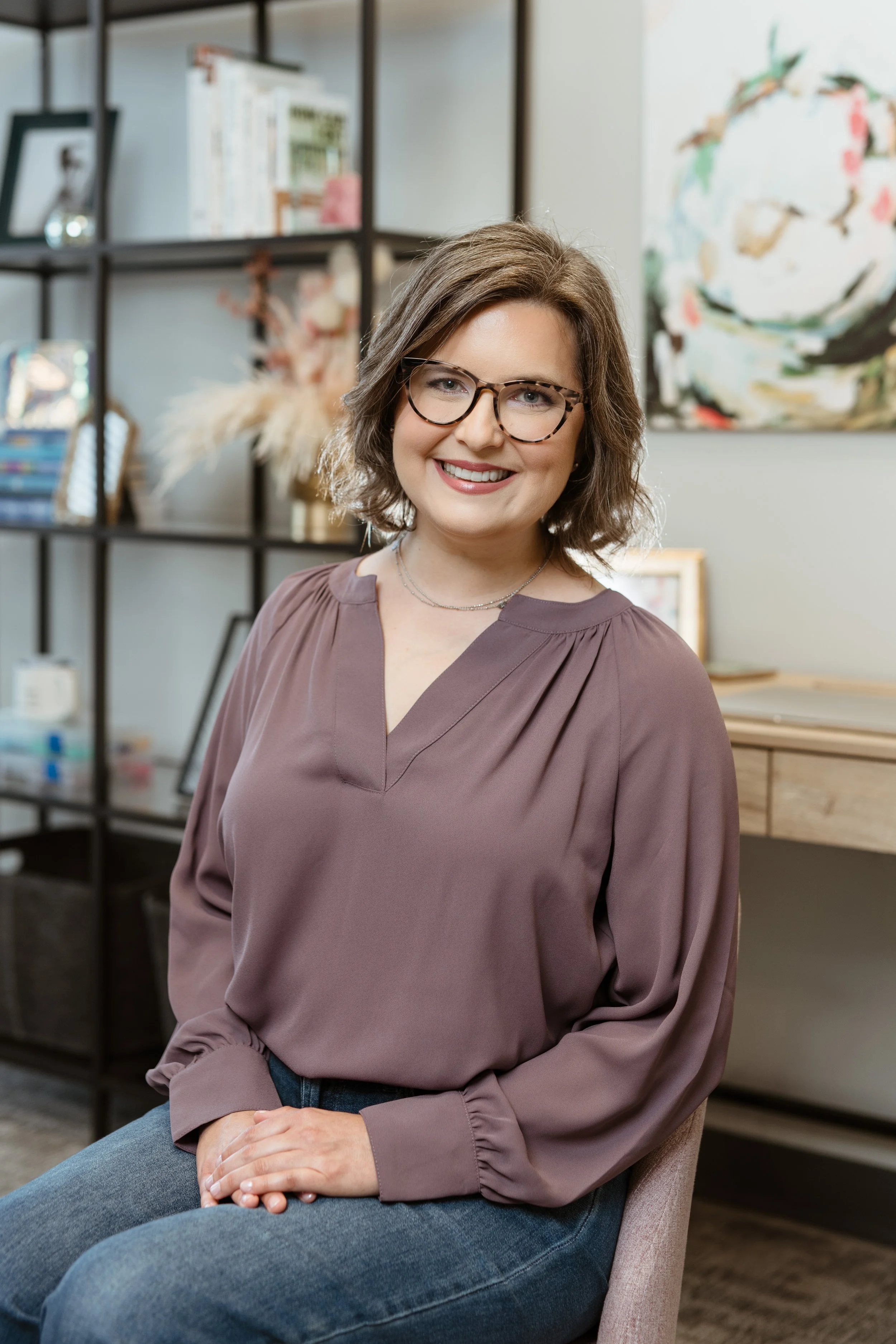 A woman with shoulder-length brown hair, wearing glasses, a mauve blouse, and blue jeans, sitting indoors with a smile.